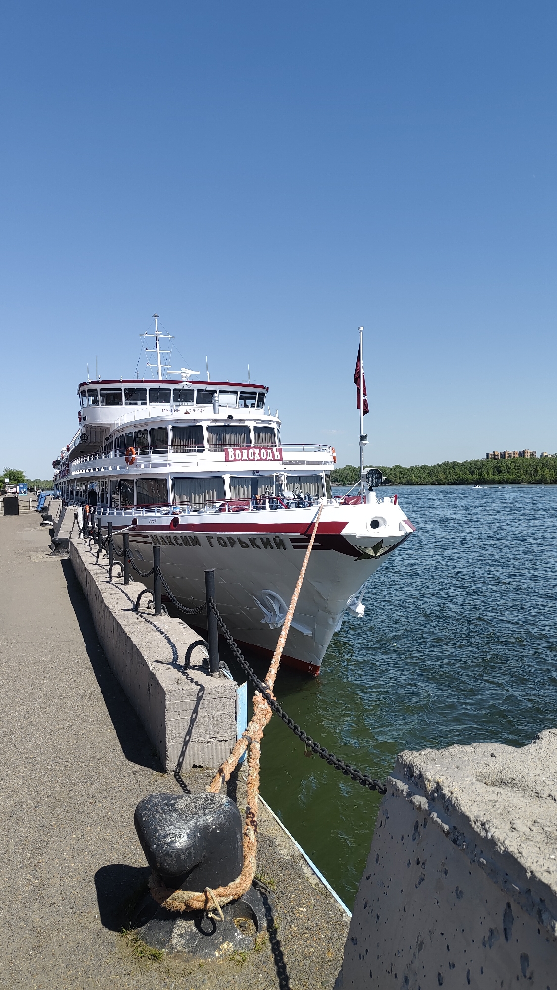 A large, white passenger ship named "МАКСИМ ГОРЬКИЙ" (Maxim Gorky) is prominently docked alongside a concrete pier under a clear, bright blue sky. The ship features multiple decks with numerous windows, and its white hull is adorned with red accent stripes. The name "Водоходъ" (Vodokhod) is also visible in red letters on the ship's superstructure, indicating its operator. A dark red flag with an emblem flies from a pole at the ship's bow. Small figures of people are visible on the ship's decks, some near the railings, suggesting passenger activity.
In the foreground, a robust black bollard is secured to the asphalt-covered pier, with a thick, worn mooring rope wrapped tightly around it, tethering the vessel to the dock. A black chain fence runs along the concrete edge of the pier. The river water is calm and appears a greenish-blue. On the opposite bank, a dense line of green trees stretches along the horizon, with some buildings visible in the distance beyond.
The scene is set on a clear, sunny day, likely during the mid-day or early afternoon, as evidenced by the intense blue sky and strong shadows cast by the pier structures. The tranquil environment suggests a peaceful stopover for the river cruise in Krasnoyarsk, Russia.