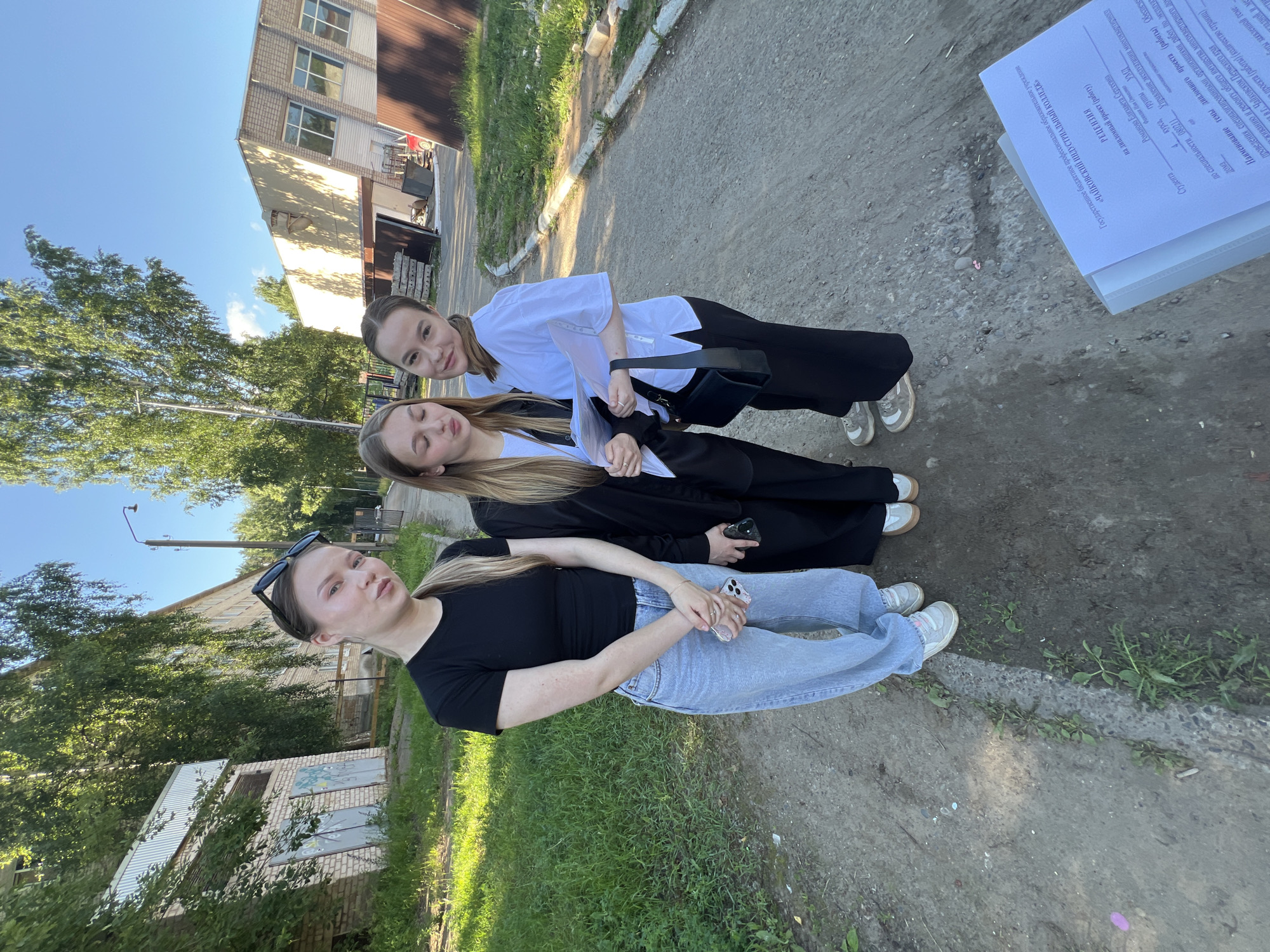 Three young women are standing on a paved walkway next to a grassy area, with buildings and trees in the background. The woman on the left is wearing a black t-shirt and light-wash jeans. The woman in the middle is wearing a black blazer over a white shirt and black pants. The woman on the right is wearing a white collared shirt and black pants. The woman on the right is holding a folder with documents. The overall setting appears to be an outdoor area associated with an educational institution, possibly a college campus, given the presence of a document labeled "Государственное бюджетное профессиональное образовательное учреждение «ЧАЙКОВСКИЙ ИНДУСТРИАЛЬНЫЙ КОЛЛЕДЖ»" (State Budgetary Professional Educational Institution "CHAIKOVSKY INDUSTRIAL COLLEGE"). The scene is illuminated by natural daylight.