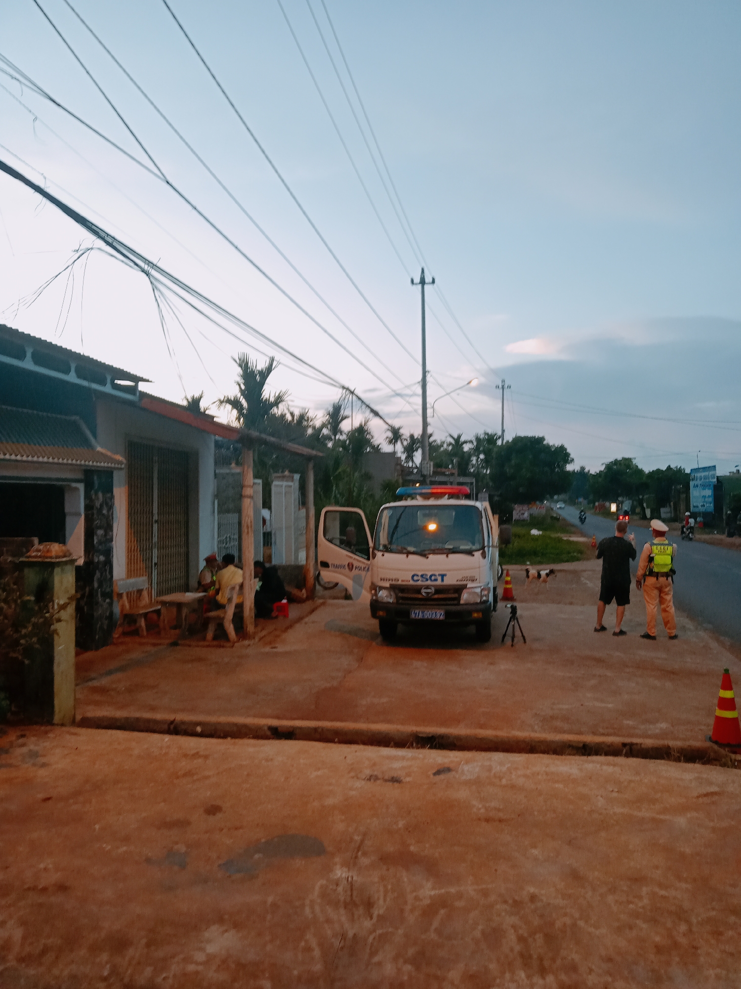 The scene is in Xuân Long, Vietnam, at twilight. A white CSGT (Traffic Police) van with flashing red and blue lights is parked partially on a sidewalk. Two men stand on the road; one in civilian clothes, and one in a police uniform. A traffic cone sits nearby. On the sidewalk adjacent to the van, several people are seated around a table in front of a building with a metal roll-up door. Overhead are numerous power lines. The road shows light traffic, including motorcycles.