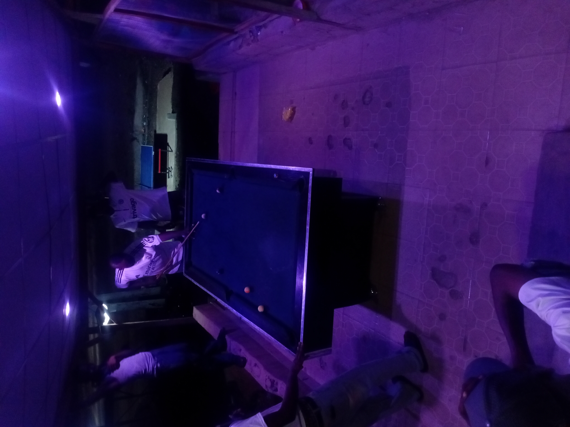 The image captures an indoor scene in Lokoja, Nigeria, illuminated by vibrant purple lighting, creating a dark, atmospheric ambiance. The central focus is a green-felt pool table where several individuals are gathered. One person, wearing a white shirt with "Emirates Fly Better" visible text, is actively playing pool, holding a cue stick over the table which has at least two balls (one white, one orange) in play.
Around the pool table, other people are present; one person in a white shirt and dark bottoms stands behind the player, while another in a white shirt is partially visible crouching or sitting in the foreground to the right. More individuals are seen sitting or standing in the background and foreground, indicating a social gathering.
The setting features walls tiled with a repeating hexagonal or octagonal pattern, strongly reflecting the purple light. The ceiling appears unfinished or rustic, with exposed beams or material. A white plastic bag bearing the text "trivago" is seen on the floor. Other objects include a blue rectangular item (possibly a speaker or equipment) and a white plastic chair in the background. The overall impression is a casual game room or bar, likely operating at night given the artificial lighting. The scene suggests a relaxed atmosphere among friends or acquaintances.