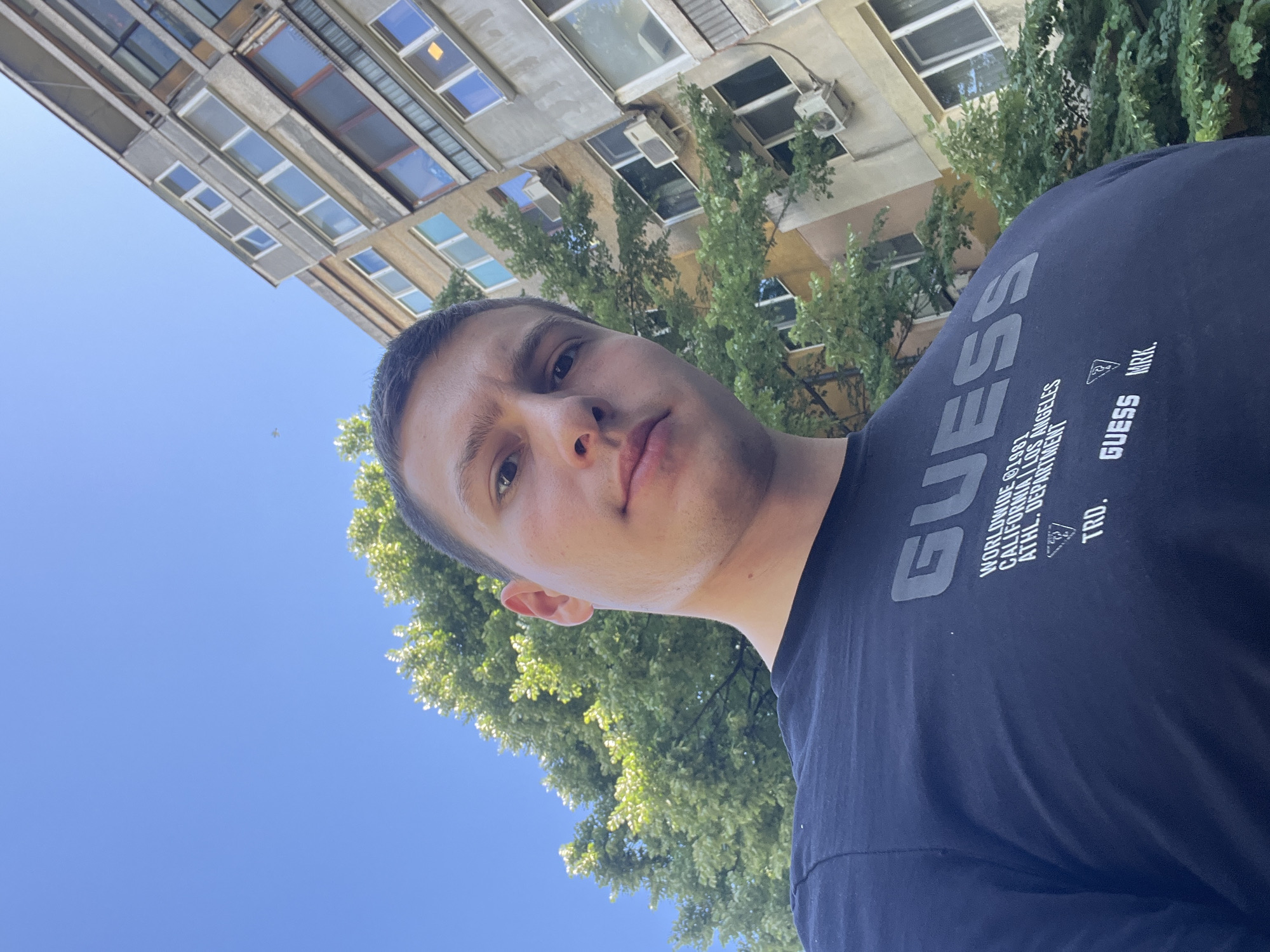 The image shows a man wearing a dark blue t-shirt with the "GUESS" logo. The man is outdoors, with a building in the background, featuring multiple windows and some air conditioning units. Green trees partially obscure the building. The sky is visible, suggesting a sunny day. The perspective is from a low angle, looking up at the man and the building. Based on the "GUESS" logo and outdoor environment, the location might be a public area in Burgas, Bulgaria, considering the question's instruction.