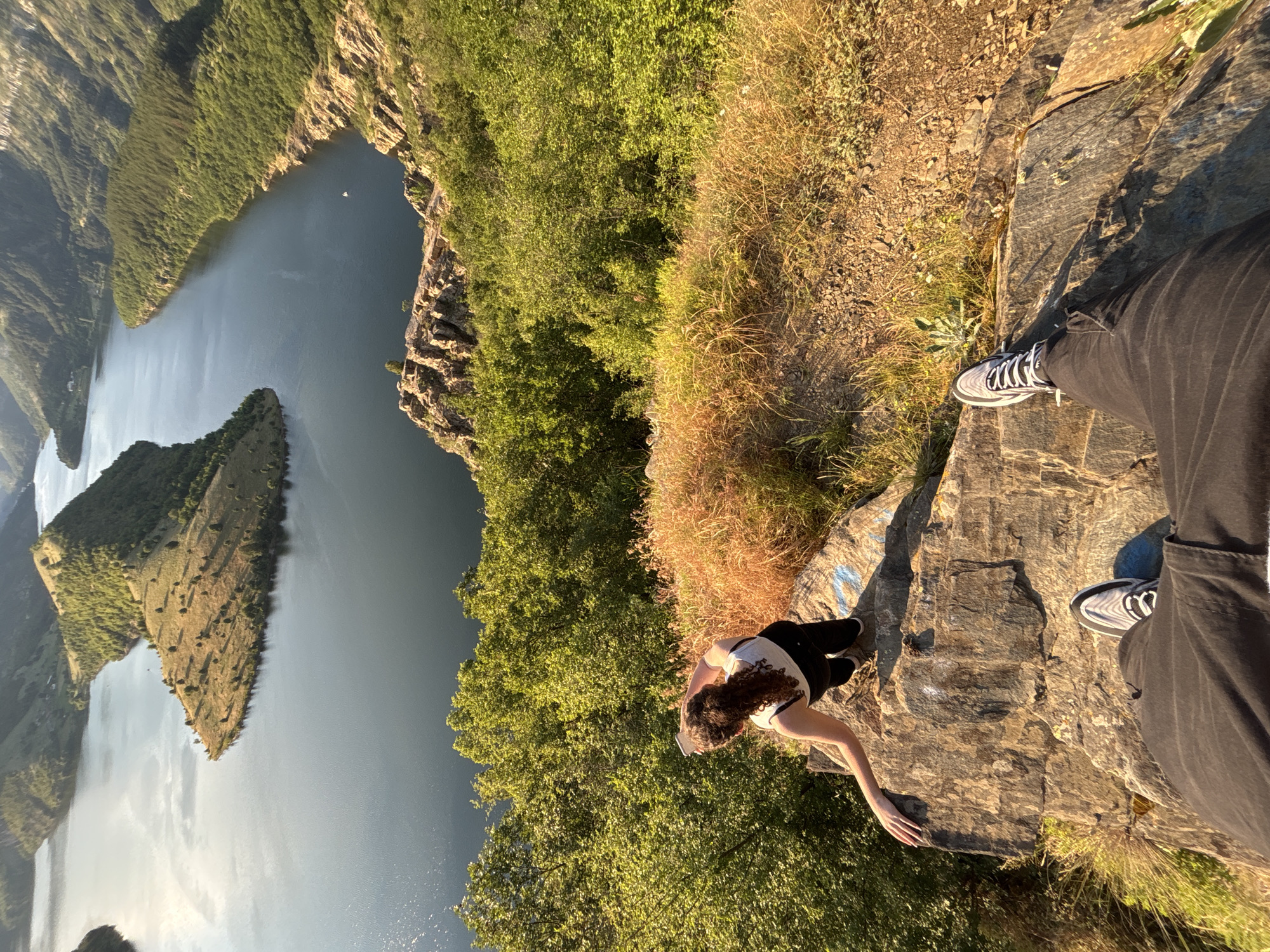 An elevated, wide-angle shot captures a young woman standing on a rocky cliff overlooking a vast, winding river and mountainous landscape. The woman, viewed from behind, is wearing a white tank top and black pants. She is holding a white mobile phone in her right hand, angled upwards. Her left arm is extended to her side, resting on the rock. The foreground is dominated by the rocky cliff edge, with the legs and feet of another person, wearing black pants and athletic shoes, visible from a first-person perspective, suggesting they are the photographer. The background features a serene body of water with a distinctive, wedge-shaped island covered in sparse vegetation and trees. The river is bordered by steep, tree-covered mountains, with the sun casting long shadows, indicating it is either early morning or late afternoon. The location is the viewpoint above the confluence of the Byala Reka and Arda rivers, near the town of Borovitsa, Bulgaria.