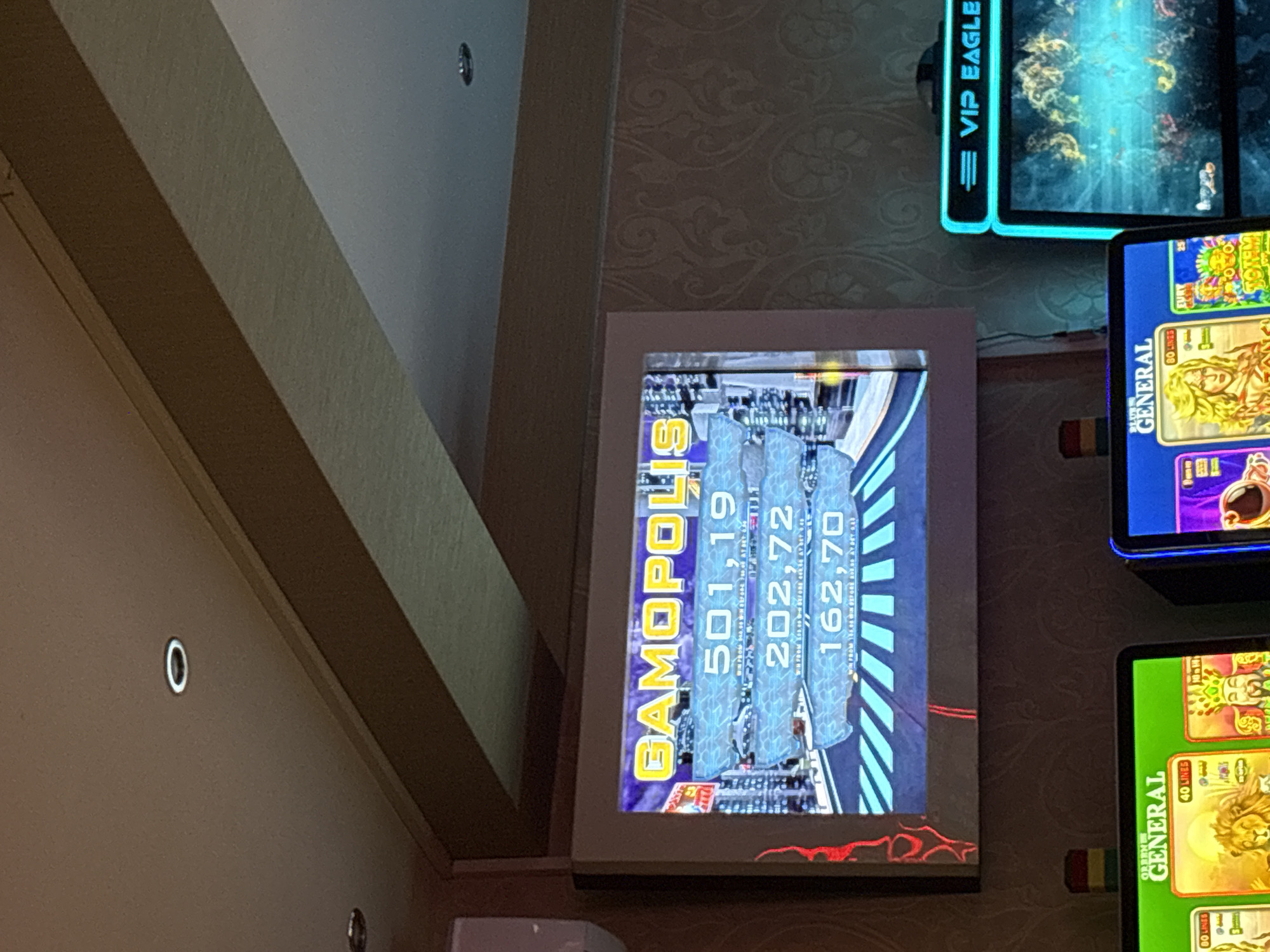 The image shows a slot machine screen displaying the text "GAMOPOLIS" at the top. Below this title, there are three numerical values displayed in separate horizontal sections: "501,19", "202,72", and "162,70". Each of these numerical displays is accompanied by smaller text which is difficult to read but appears to be related to wins or bets. The background of the screen features a stylized cityscape. To the right of this screen, another slot machine with a glowing blue frame is partially visible, displaying the text "VIP EAGLE". Below the "GAMOPOLIS" screen, parts of two other slot machine screens are visible. The left one has a green background and displays "GENERAL" along with imagery of a lion and the text "40 LINES". The right one has a purple background and displays "GENERAL" with imagery of a person. The environment appears to be an indoor establishment, possibly a casino or a gaming hall. The ceiling features recessed lighting. The wallpaper has a patterned design. The image does not contain any information or visual cues that would allow for the identification of the city of Karlovo, Bulgaria.