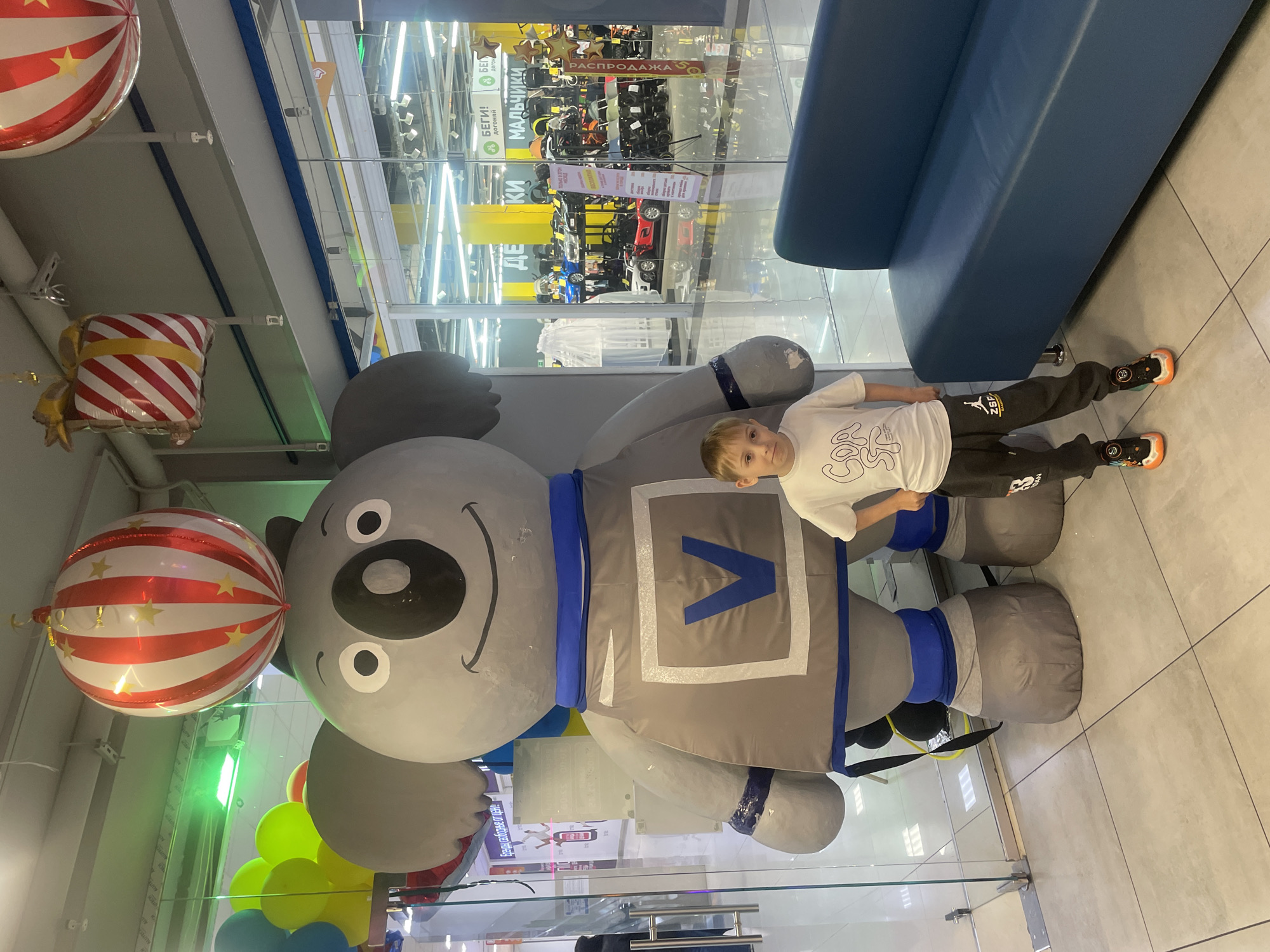 A boy is standing in a building in Sochi, Russia, next to a large stuffed gray koala bear with blue trim. The koala has a large "V" on its chest. Several red and white balloons are visible, as well as other toys and merchandise behind a glass wall. A blue couch sits adjacent to the boy.