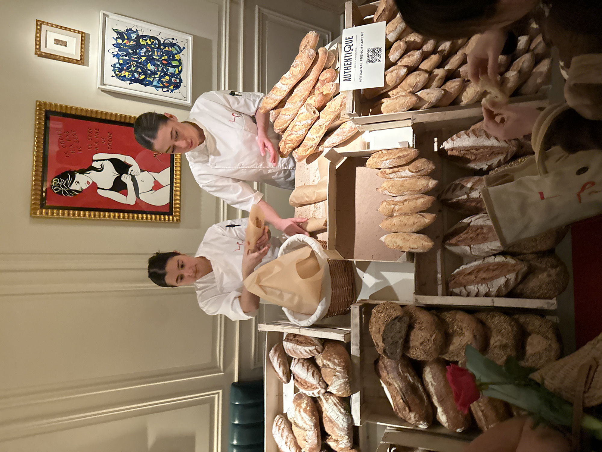 This image captures an artisanal French bakery display within an upscale interior, located in London, United Kingdom. Two women in white chef's jackets, both featuring an "LP" emblem, are positioned behind a comprehensive bread arrangement. The woman on the left is seen placing a loaf into a brown paper bag held within a wicker basket, while the woman on the right stands with her hands near a stack of baguettes, overseeing the selection.
The extensive bread display is presented in several wooden crates. Prominent items include numerous baguettes, stacked both horizontally and vertically. Other bread types visible are round loaves (boules) and oblong loaves with decorative scoring, some exhibiting darker crusts indicative of whole grain or sourdough. A white sign on the right-hand display crate reads "AUTHENTIQUE BOULANGERIE ARTISANAL FRENCH BAKERY" and includes a QR code.
In the lower right foreground, a person's hands are visible, one reaching for or holding a piece of bread. A single red rose with a green stem is positioned in the lower left foreground.
The background features a light cream-colored wall with classic paneling. Three framed artworks are mounted on this wall. The largest, centrally located, is a gold-framed piece depicting a stylized woman in a black bra and red panties with a red heart, set against a red background. French text on this artwork reads "LE COEUR N'A PAS DE SEXE / LE SEXE N'A PAS DE COEUR." To its right, a smaller gold-framed abstract piece and a white-framed abstract painting with blue, yellow, and black curvilinear patterns are displayed. A section of a dark green upholstered booth or bench is partially visible on the far left. The scene is illuminated by warm, ambient lighting.