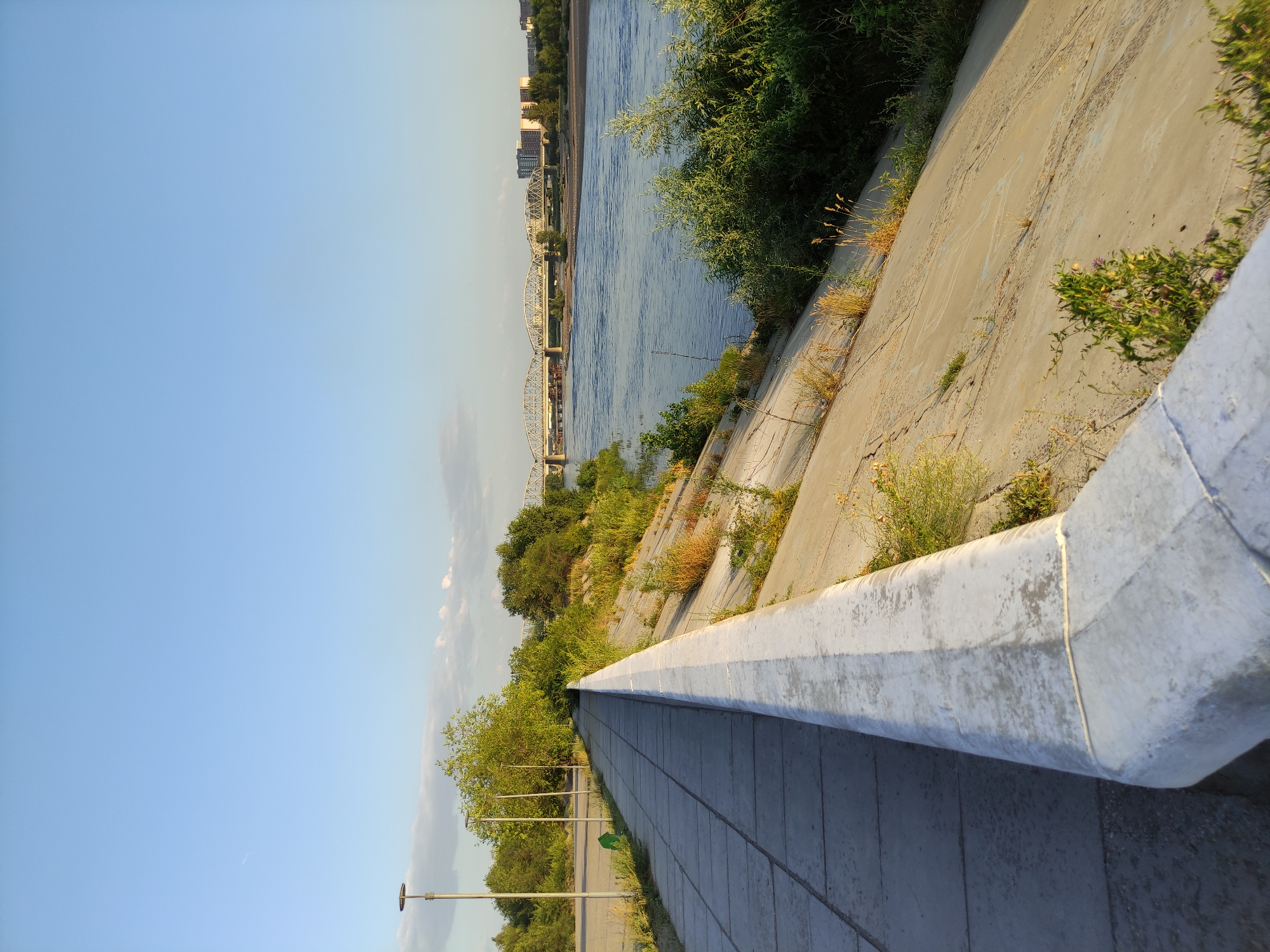The image depicts a concrete embankment along the Irtysh River in Semey, Kazakhstan. A paved walkway runs alongside the embankment, leading the viewer's eye towards a distant bridge with multiple arches spanning the river. Lush green trees and vegetation border the riverbank, contrasting with the man-made concrete structures. The sky is a clear, pale blue, indicating fair weather, and the scene is bathed in the warm light of late afternoon or early morning. There are no people visible in the image. The overall impression is one of a serene, urban waterfront area.