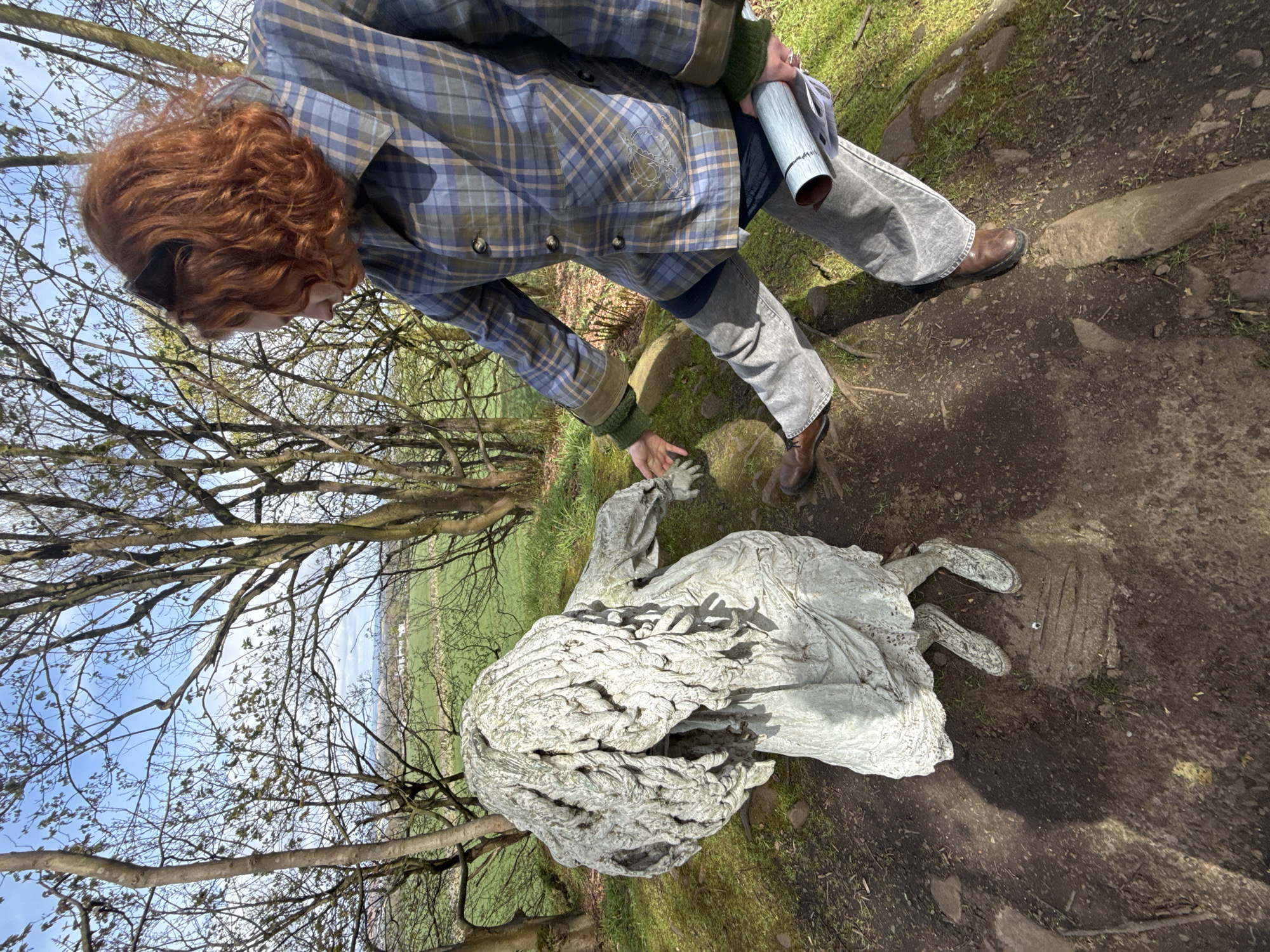 A person with curly red hair, dressed in a blue and green plaid jacket, green sweater sleeves, grey jeans, and brown boots, leans down towards a light-colored statue lying on the ground. The statue appears to be a human-like figure with textured, flowing hair or drapery. The person's right hand is extended towards the statue's hand, suggesting an interaction or observation. They hold a rolled-up item, possibly a map or document, in their left arm.
The scene is an outdoor setting under bright daylight, with clear shadows indicating sunshine. The ground consists of a dirt path mixed with patches of grass and scattered rocks. In the background, deciduous trees with mostly bare branches or very early foliage stand against a clear blue sky. Beyond the trees, vibrant green fields extend into the distance. The setting suggests a natural environment, possibly a park or rural trail, during early spring. There is no visible text in the image.