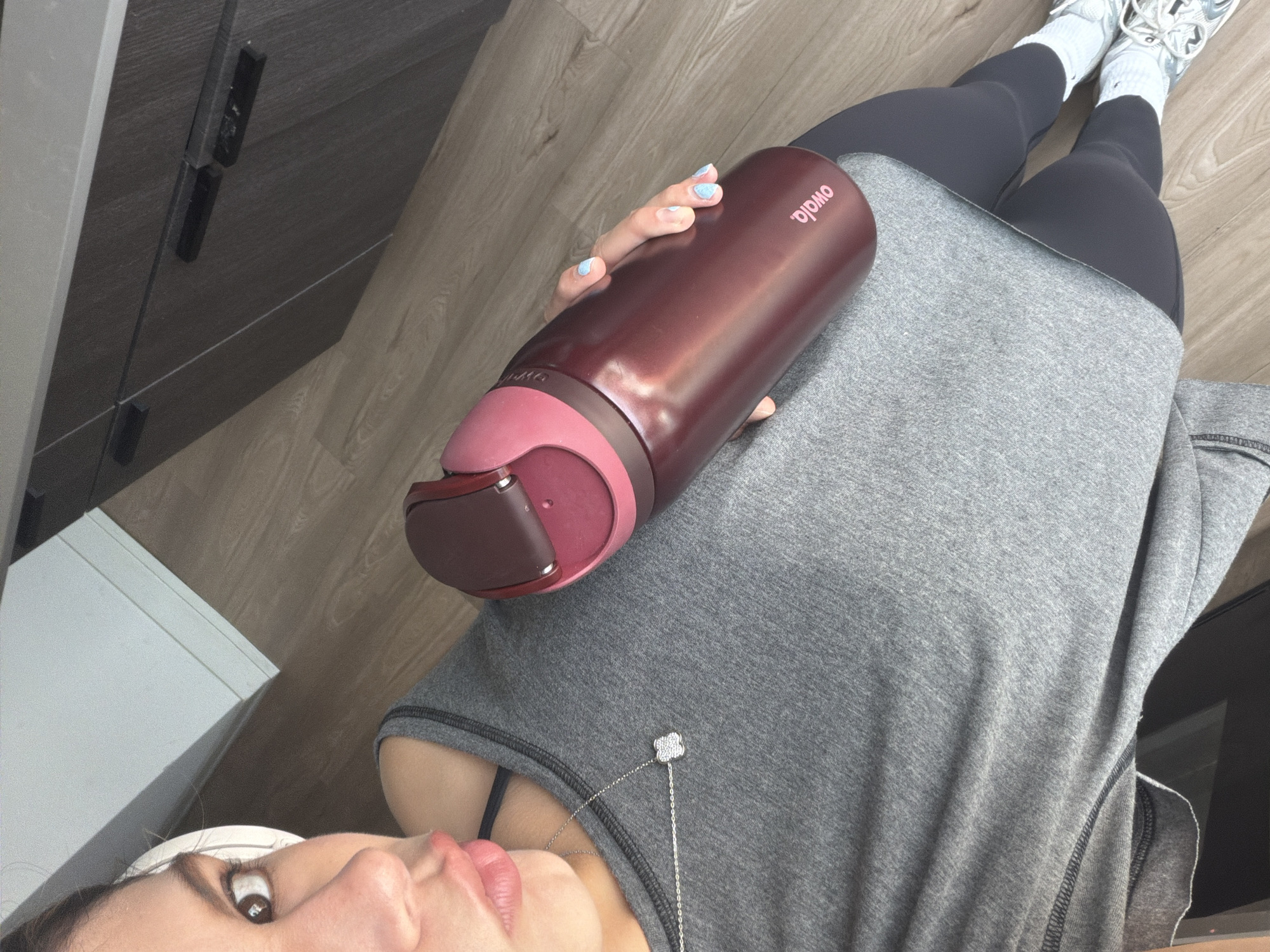 A person is lying down, possibly on the floor, holding a reddish-brown Owala brand water bottle. They are wearing gray leggings, white sneakers, and a gray t-shirt. A silver necklace with a four-leaf clover pendant is visible around their neck. The background shows a wooden floor and a dark-colored cabinet. The location is likely indoors in Austin, United States of America, but the lack of specific landmarks prevents precise identification.