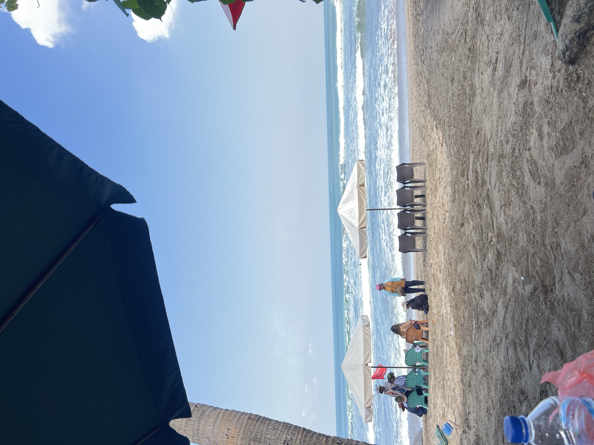 The image depicts a sandy beach scene in Kuta, Indonesia, on a clear, sunny day. In the foreground, a blue-capped water bottle and a red plastic bag are visible on the sand. The middle ground features a row of empty plastic chairs beneath two large umbrellas, with a few people gathered near the left umbrella. One individual, wearing a yellow hat and orange shirt, stands by the umbrella, while others are seated or standing nearby. The ocean stretches out in the background, with moderate waves breaking on the shore. The sky is a vibrant blue with scattered white clouds. A portion of a palm tree trunk is visible on the left side of the frame, and a large dark green umbrella occupies the top portion of the image, casting a shadow.
