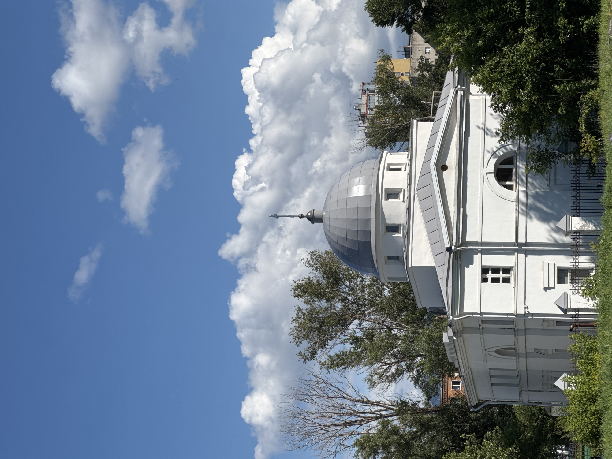 The image shows a white, classical building with a grey dome, likely a church or a mosque, set against a bright blue sky with dramatic white clouds. The building has a low-pitched roof and several windows, some arched. The dome is topped with a cross. Lush green trees surround the building, with a few bare, skeletal trees visible to the left. In the background, there are other buildings, including a taller, yellow building. A black metal fence runs along the front of the property, separating it from a strip of green grass and vegetation. The lighting suggests it is daytime, with strong sunlight casting shadows. The architectural style and the presence of trees suggest an urban or suburban setting. This building is located in Nizhny Novgorod, Russia, and is likely the Astrakhanskaya Mosque.