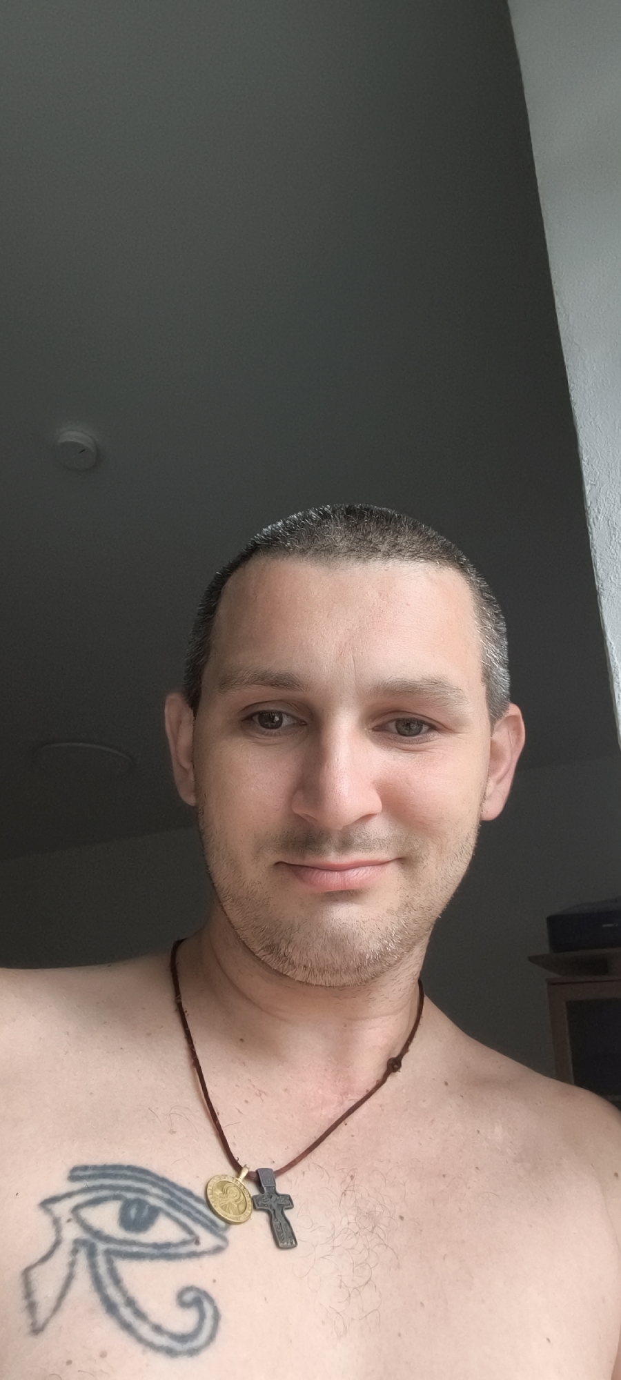 The image shows a close-up of a man's face and chest. He has short, dark hair and light brown eyes. He is wearing a necklace with two pendants: a gold-colored oval pendant and a dark-colored cross pendant. On his chest, to the left, there is a tattoo of an eye. The man is shirtless, and his skin is fair with some light hair on his chest. The background appears to be indoors, with a dark wall and a lighter wall or window to the right. There is a ceiling light fixture visible in the upper left corner. The image is cropped vertically, with the top of the man's head and shoulders visible.
There is no information in the image that indicates a specific location in the city of Wolgast, Germany.