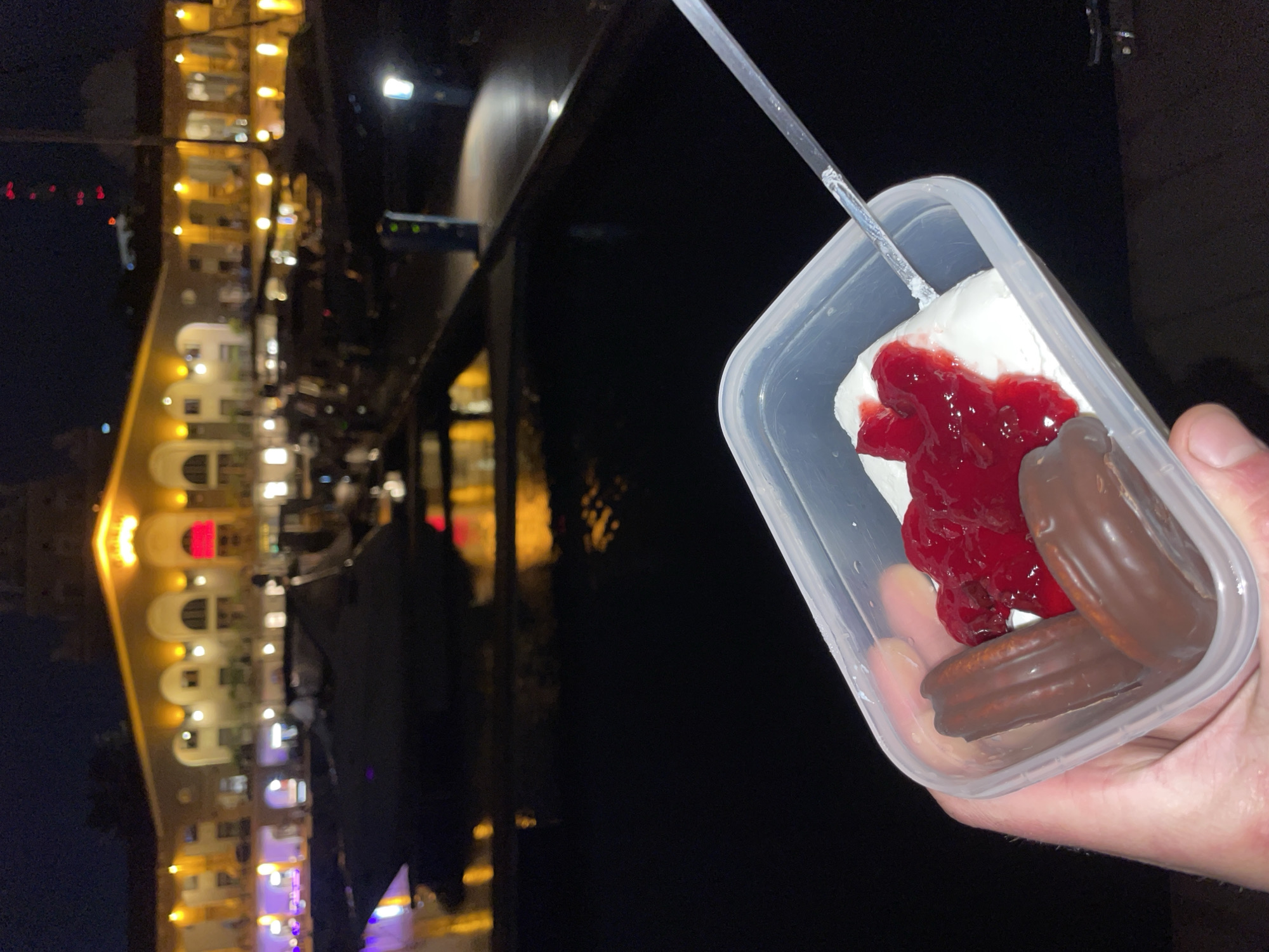 A hand holds a clear plastic container filled with food, specifically two chocolate-covered cookies, white cream, and red cherry filling. A clear plastic spoon rests inside the container. The background shows the night sky and a building lit up with yellow and red lights, which is reflected in the water. Based on the architectural style and lighting, this is likely a view in Sochi, Russia.