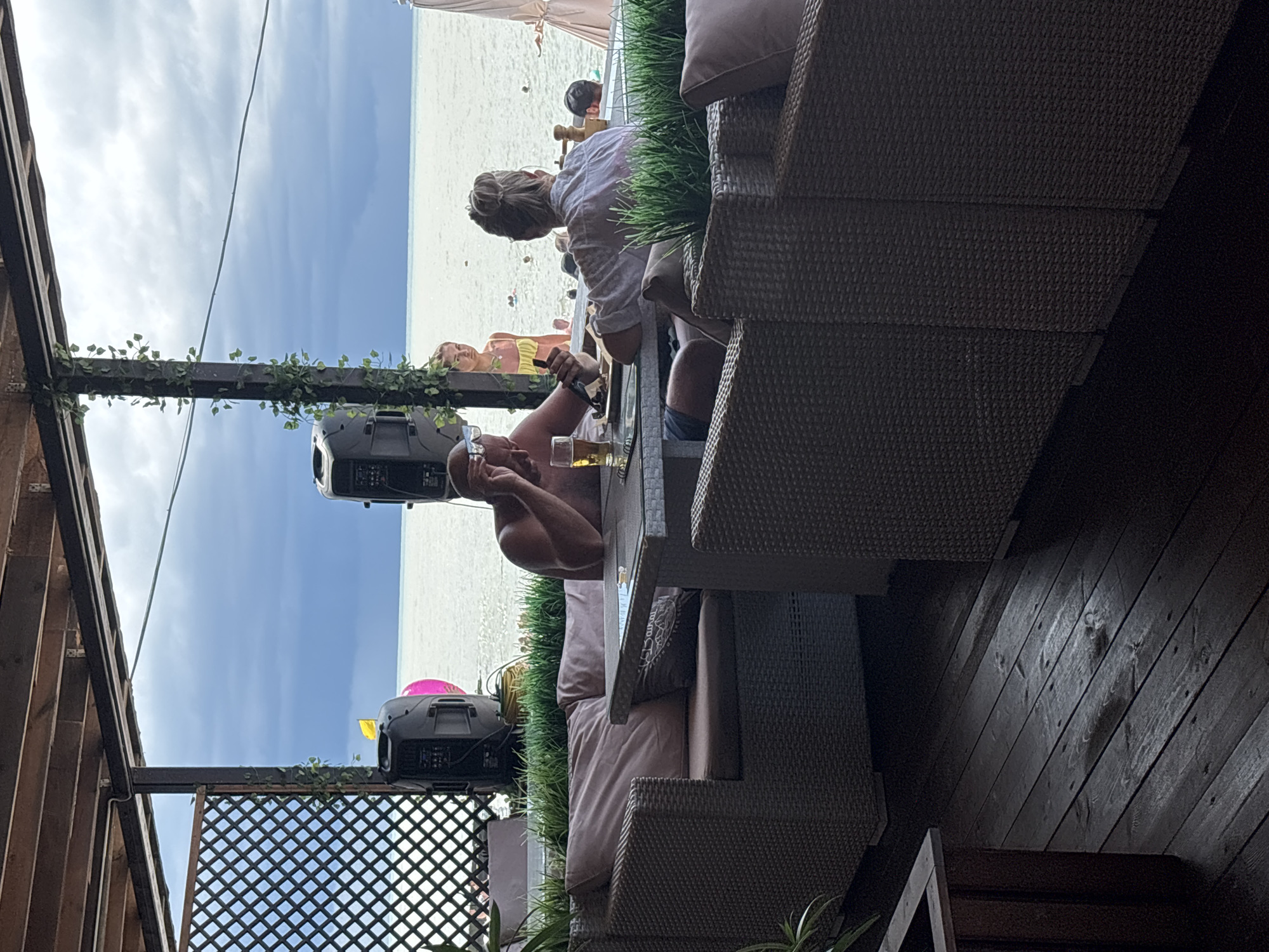 This image shows a view from an outdoor cafe with a thatched roof overlooking the Black Sea in Sochi, Russia. In the foreground, two people are seated at a table. A man with a bald head is on the left, wearing blue shorts and holding a glass of beer and a plate of food. He is looking down at the food. A woman with blonde hair tied up in a bun is on the right, wearing a white shirt and looking away from the camera. She is also seated at the table.
In the background, a young woman in a yellow bikini stands near the water. Several other people are visible in the sea, some swimming and others floating. Two large black speakers are positioned on the left side of the frame, near a latticed screen. A pink inflatable object is also visible near the speakers. The sky is cloudy. The cafe features wicker furniture and wooden decking.