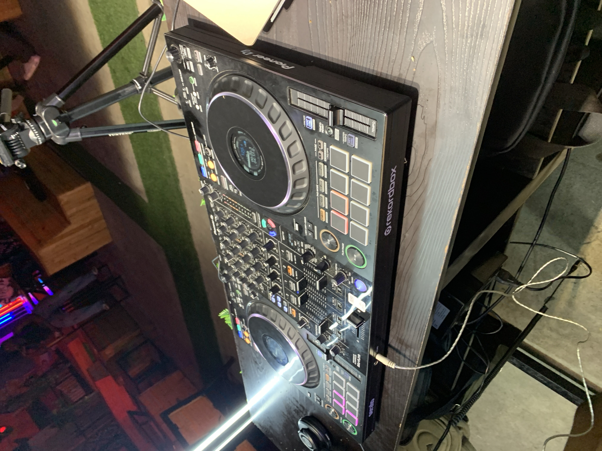 An eye-level, medium shot captures a Pioneer DJ controller on a wooden table in what appears to be an outdoor or semi-outdoor venue at night. The controller, a Pioneer DJ DDJ-FLX10, is black with illuminated buttons and jog wheels. A tripod with a camera is visible in the upper right corner, behind the DJ equipment. In the background, there are tables and chairs, some with people sitting, illuminated by colorful neon lights. The overall scene suggests a nightlife or event setting.