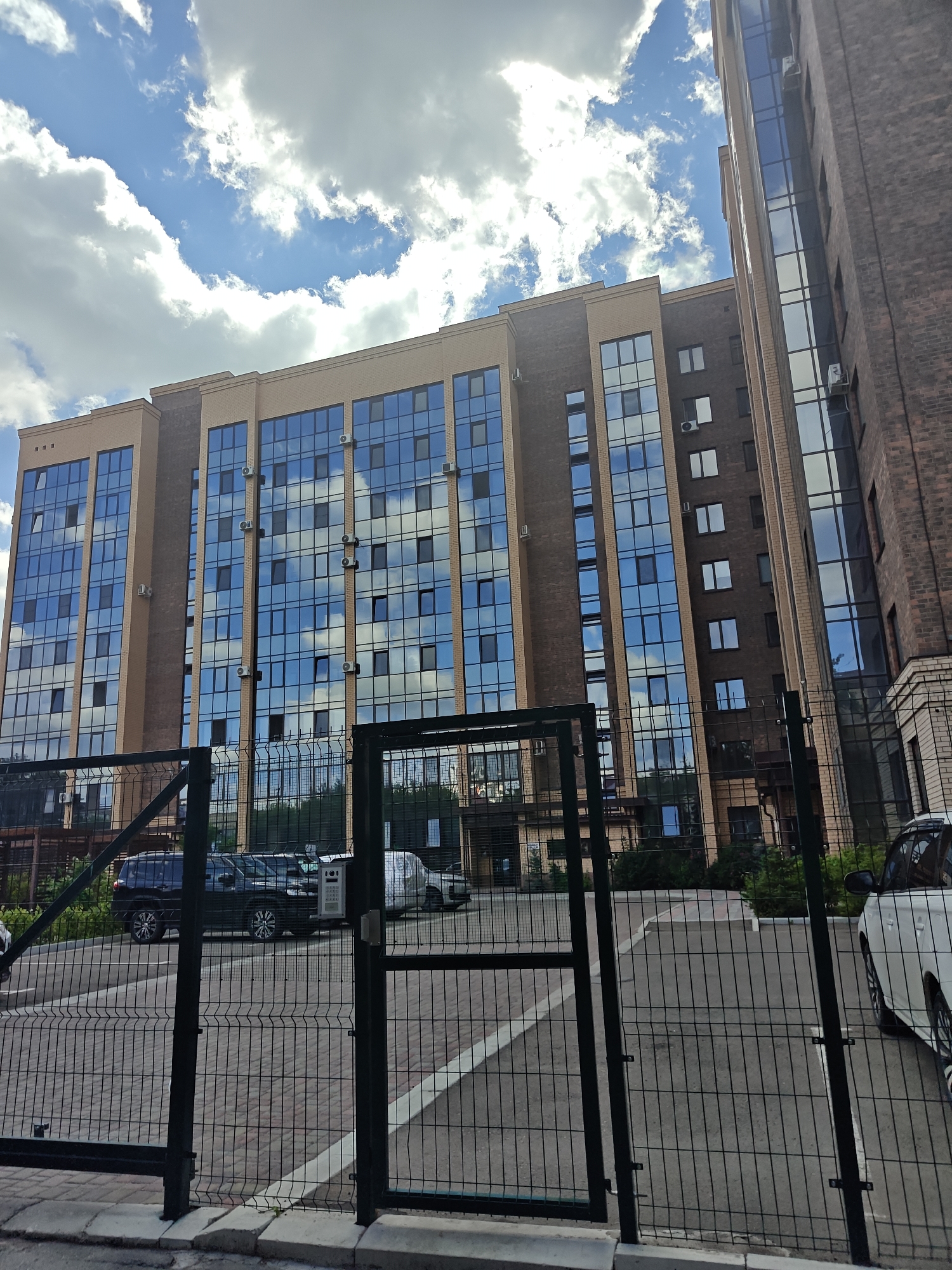 A low-angle shot captures a modern, multi-story building with a facade of beige brick and large, blue-tinted glass windows reflecting a cloudy sky. A dark brick building stands to the right, also featuring extensive glass panels. In the foreground, a black metal fence with a gate partially obscures a parking lot. Several vehicles, including a black SUV, a white van, and a white car, are visible in the lot. The sky above is a vibrant blue with scattered white cumulus clouds. The scene is likely a residential or commercial complex in Petropavl, Kazakhstan, given the architectural style and the presence of parked vehicles.