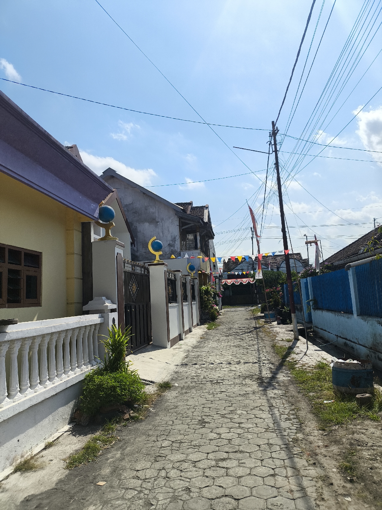 The image shows a narrow, paved alleyway in a residential area in Lumajang, Indonesia. The alley is flanked by houses on both sides, with a blue fence on the right and a white balustrade on the left. The houses have a traditional architectural style with tiled roofs. Colorful triangular flags and pennants are strung across the alley, suggesting a celebration or festival. A utility pole with numerous wires stands in the middle of the alley. The ground is paved with hexagonal stones. The sky is blue with a few white clouds. There are no people visible in the image.