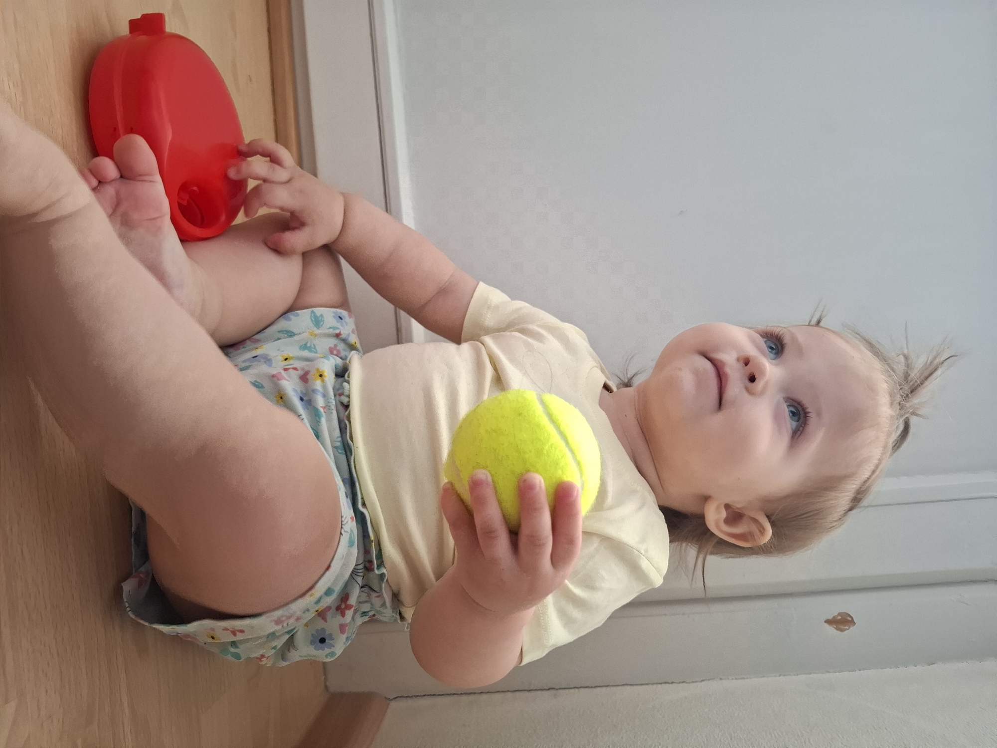 A baby is lying on a wooden floor, holding a tennis ball in their right hand. The baby is wearing a yellow t-shirt and a diaper with a floral pattern. A red plastic toy sits next to the baby's left leg. The background shows a white wall. The location is Chaikovsky, Russia.