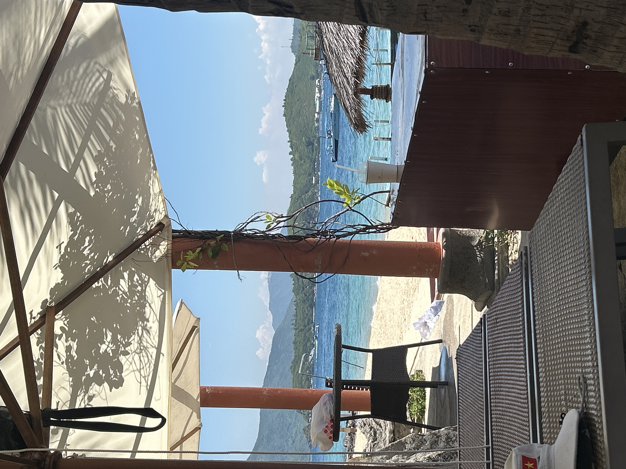 This media file depicts a tropical beach scene, likely in Bac Nha Trang Ward, Vietnam. The foreground is dominated by the structure of an open beach umbrella, casting dappled shadows from palm fronds onto its white fabric. The umbrella structure is made of light-colored wood with dark straps. To the right, a portion of a table with a textured surface is visible, along with a chair.
Beyond the immediate foreground, a view opens up to a vibrant blue ocean stretching towards a verdant, mountainous coastline. Several boats are anchored on the water, suggesting a leisurely atmosphere. The beach itself is a strip of light sand. A thatched-roof structure is visible on the right side of the scene, further contributing to the tropical ambiance. A clear plastic cup with a straw sits on a surface, hinting at refreshments.
The time of day appears to be daytime, with bright sunlight casting distinct shadows. The sky is a clear, bright blue, indicating fair weather. The overall mood is relaxed and inviting, evoking a sense of vacation and seaside leisure. There is no visible text in the image.