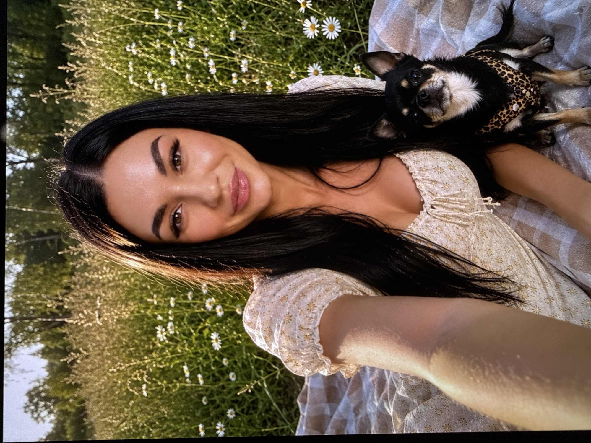 In the city of Letniy Otdykh, Russia, a woman and her small black and tan Chihuahua dog are posing for a selfie on a picnic blanket. The woman has long, dark hair and is wearing a floral-print dress. The dog is wearing a leopard-print harness. They are in a field of tall grass and white wildflowers, with trees visible in the background. The sun is shining, casting a warm glow on the scene.