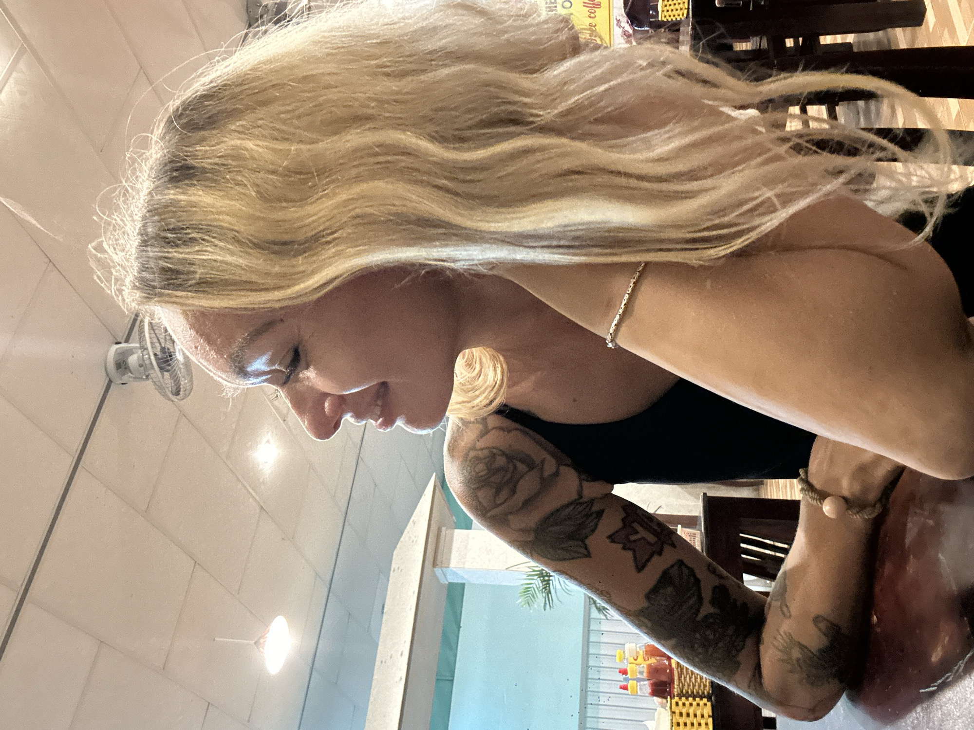 A medium shot captures a woman with blonde hair in profile, seated at a wooden table. She is looking to her left with a slight smile, her left arm resting on the table. Her arm is adorned with tattoos, including roses and other designs. She is wearing a black tank top and a thin bracelet on her left wrist. The background suggests an indoor setting, possibly a restaurant or cafe, with a ceiling fan, lighting fixtures, and a portion of a white structure with a green background visible. In the distance, on the table, are three condiment bottles and a yellow woven basket. The overall lighting is warm and somewhat dim, indicating it might be daytime but not overly bright. The context of Phan Thiết, Vietnam, suggests a potentially humid and warm climate, which could influence the overall atmosphere of the scene.