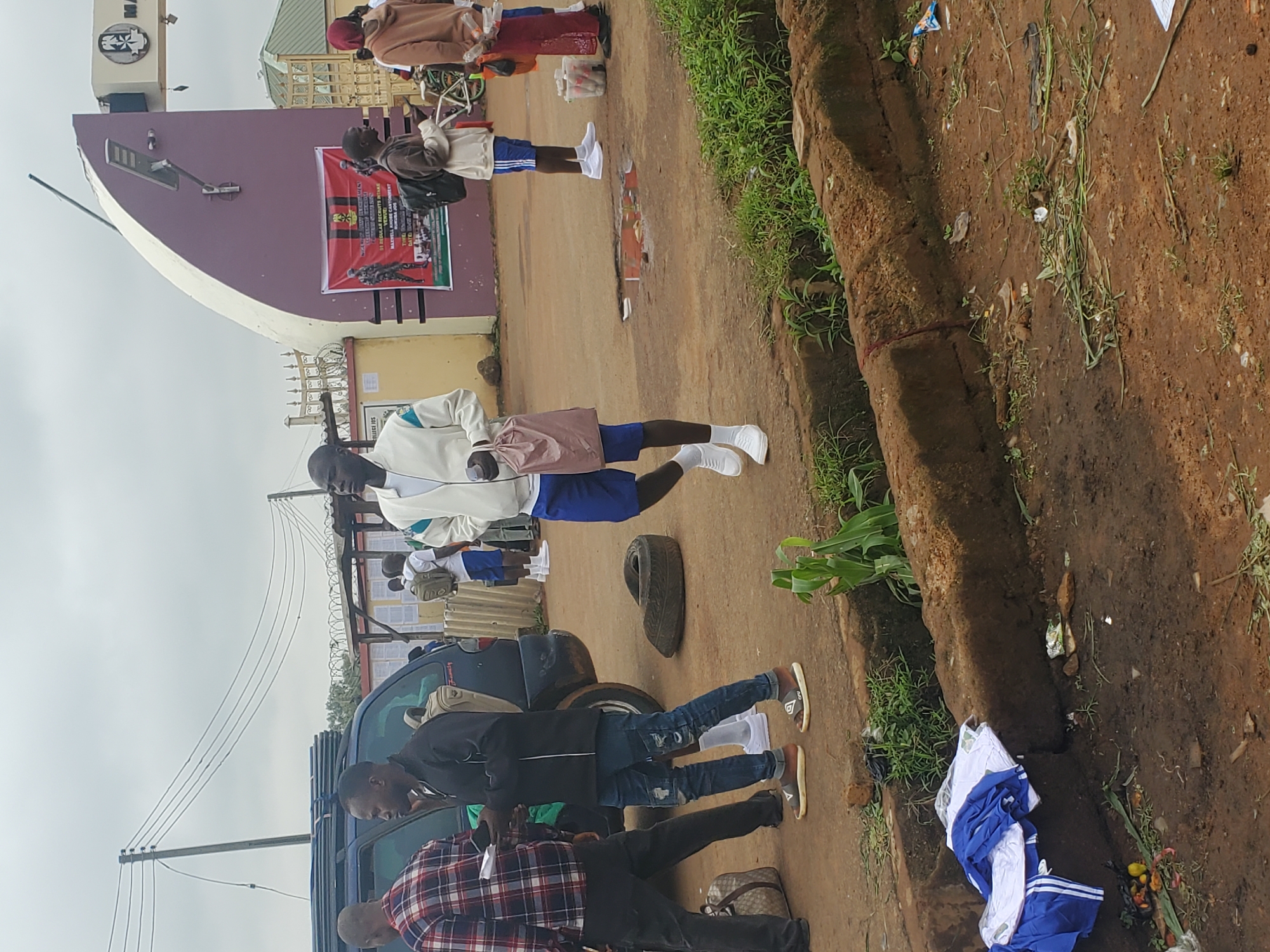 The image depicts the exterior of a building with a large, curved, purple archway, likely an entrance to a facility. A banner advertising "NIGERIAN ARMY RECRUITMENT MASSIVE INTAKE" is displayed on the archway. Several individuals, appearing to be recruits or applicants, are present outside. One man in a white jacket and blue shorts walks towards the camera carrying a pink bag. Another man in a black jacket and blue jeans stands near a dark-colored SUV. Other individuals, some carrying backpacks and wearing athletic attire, are visible in the background. The ground is muddy and uneven, with debris and vegetation scattered around. The scene appears to be outdoors, possibly in a public area or near a military installation in Bahit, Nigeria.