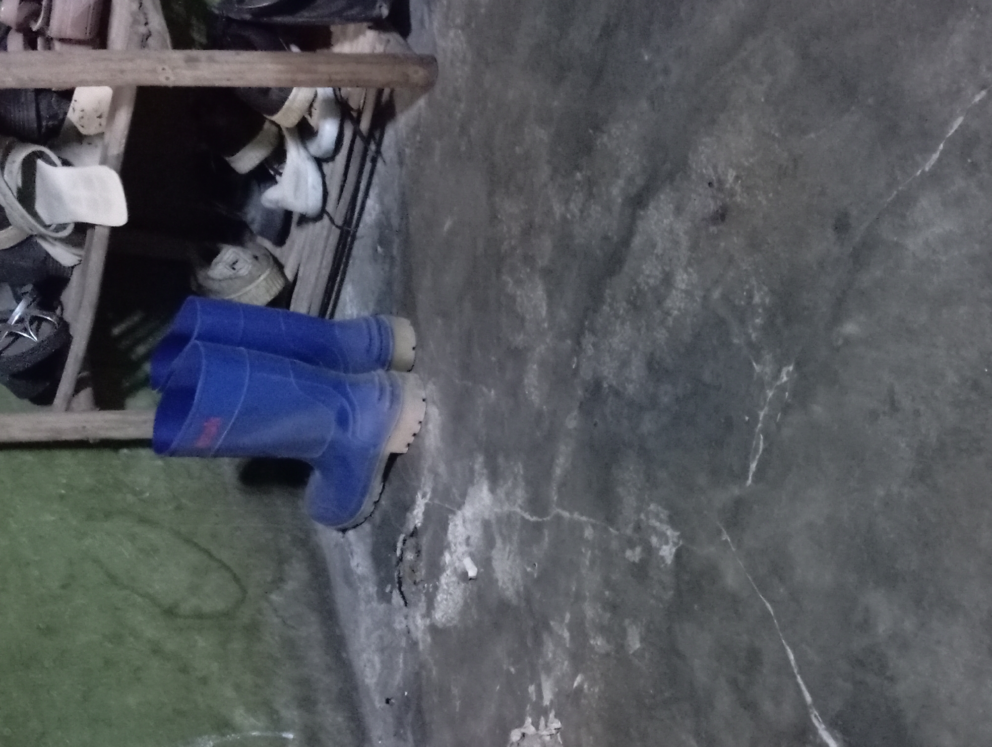 The image depicts a dimly lit corner of a room, likely in Rejosari, Indonesia. In the foreground, a pair of bright blue rubber boots stands upright on a cracked, grey concrete floor. To the right, a wooden shelf unit is visible, filled with various shoes. Some of these appear to be sneakers and casual shoes, in colors like white, black, and beige. The wall behind the boots has a textured, green surface with what appears to be a faint, hand-drawn outline. The lighting suggests an indoor environment, possibly a storage area or a less-used space. No people are visible in the frame, and no specific actions are taking place, other than the boots standing ready for use.