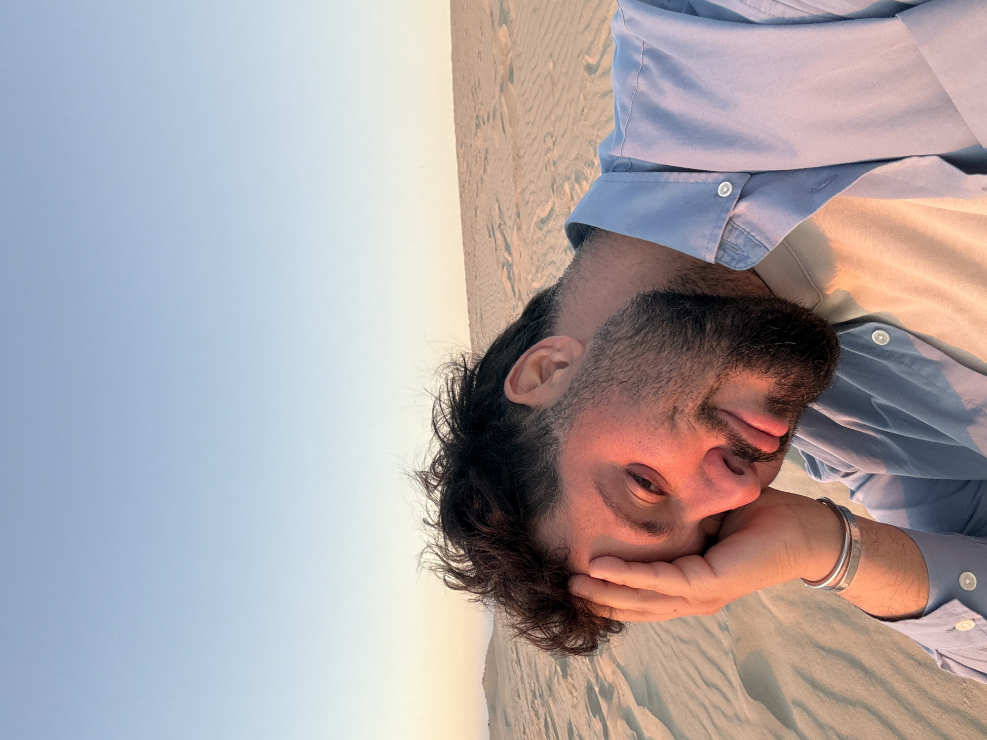 A man is taking a selfie in the sand dunes of La Teste-de-Buch, France. The man has dark, messy hair and a beard, and is wearing a light blue button-down shirt over a beige t-shirt. His left hand is resting on his forehead, and he is looking at the camera with a slight smile. The sand dunes stretch out behind him, with a clear blue sky above. The lighting suggests it is either sunrise or sunset.