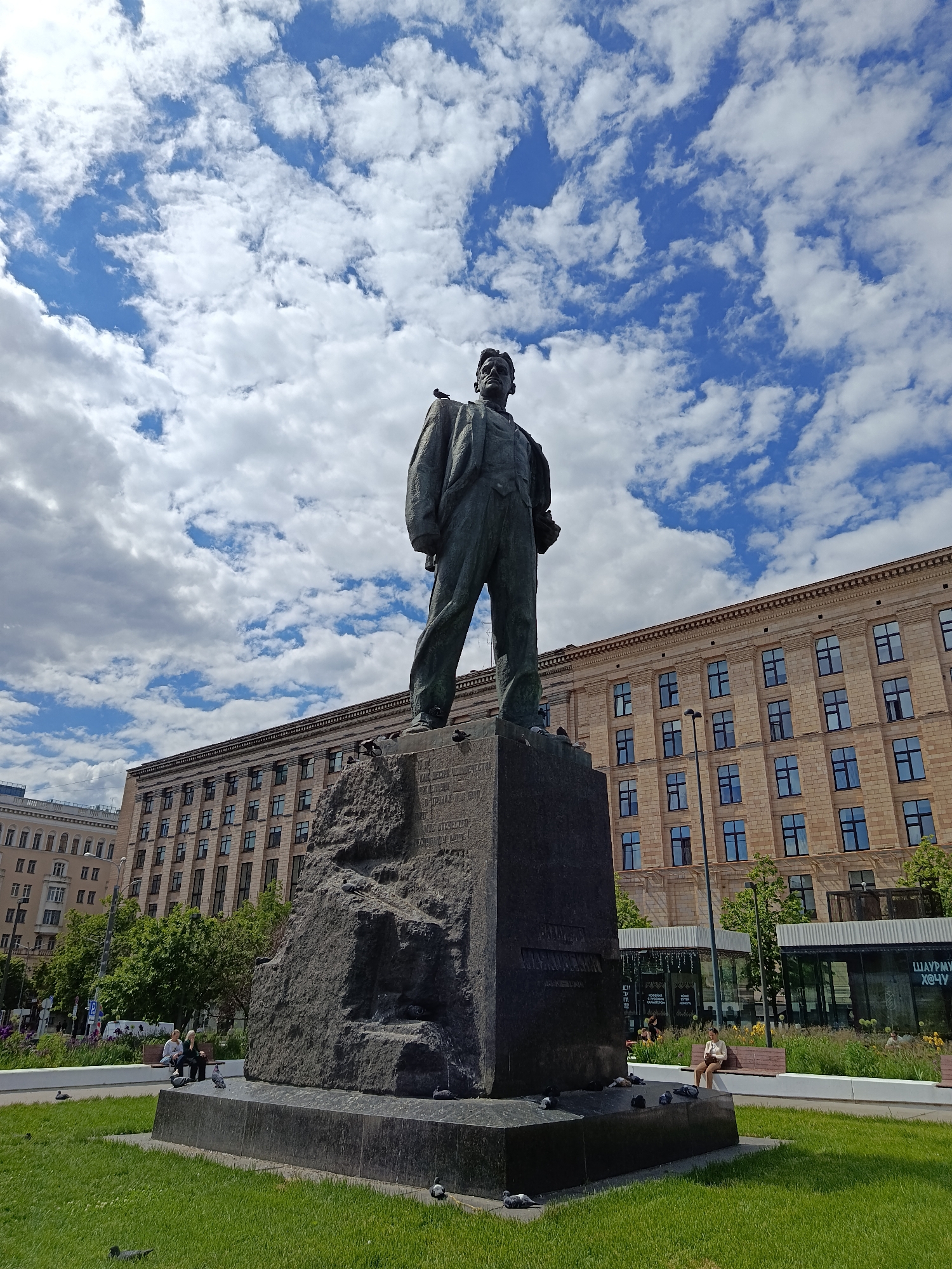 The image displays a large, dark-colored statue of a man in a suit, standing on a prominent, rough-hewn stone base, which rests on a darker plinth. The statue depicts the man looking slightly upwards and to his left. Pigeons are visible perching on the statue's shoulders, head, and base, as well as on the surrounding green grass.
In the middle ground, a long, multi-story building with a light-colored facade and numerous large windows stretches across the scene. Its design features classical architectural elements, such as cornices. To the left, another multi-story building with a lighter facade is partially visible. To the right of the statue, behind the grassy area, modern kiosks with expansive glass windows and outdoor seating areas are present.
The sky above is a vibrant blue, filled with scattered white cumulus clouds, indicating a partly cloudy but bright daytime. The foreground consists of a meticulously maintained green lawn surrounding the statue. Several people are visible: two individuals are seated on a bench to the left of the statue, and another person is seated on a bench to its right, near the modern kiosks.
Notable details include the presence of numerous pigeons on the statue and the ground, and the bright, sunny weather with clear skies despite the clouds. Visible Cyrillic text on the rough stone base of the statue is largely indistinct. However, text on a modern kiosk to the far right is discernible as "ШАУРМУ ХОЧУ" (SHAURMU KHOCHU), which translates to "I want shawarma." The scene captures a tranquil moment in a public space.