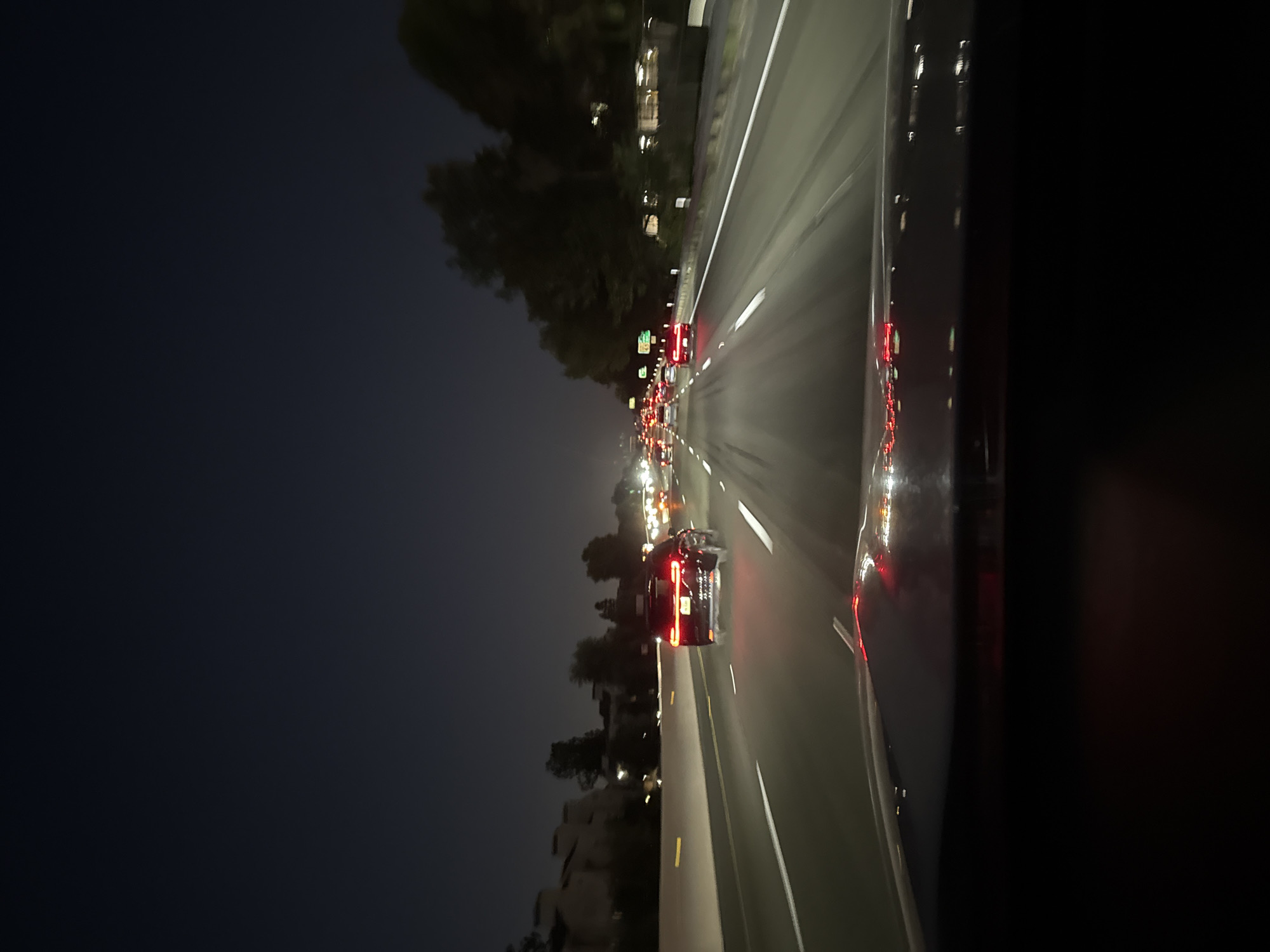 The image depicts a nighttime view from the perspective of a vehicle traveling on a multi-lane highway in Los Angeles, California. The sky is dark blue, indicating it is dusk or night. The highway is well-lit by streetlights and the headlights and taillights of other vehicles. Several cars are visible on the highway, traveling in the same direction as the viewer's vehicle. In the foreground, the hood of the viewer's car is visible, with its brake lights illuminated. On the left side of the highway, buildings and trees can be seen, suggesting an urban or suburban environment. The overall scene is one of typical highway traffic at night.