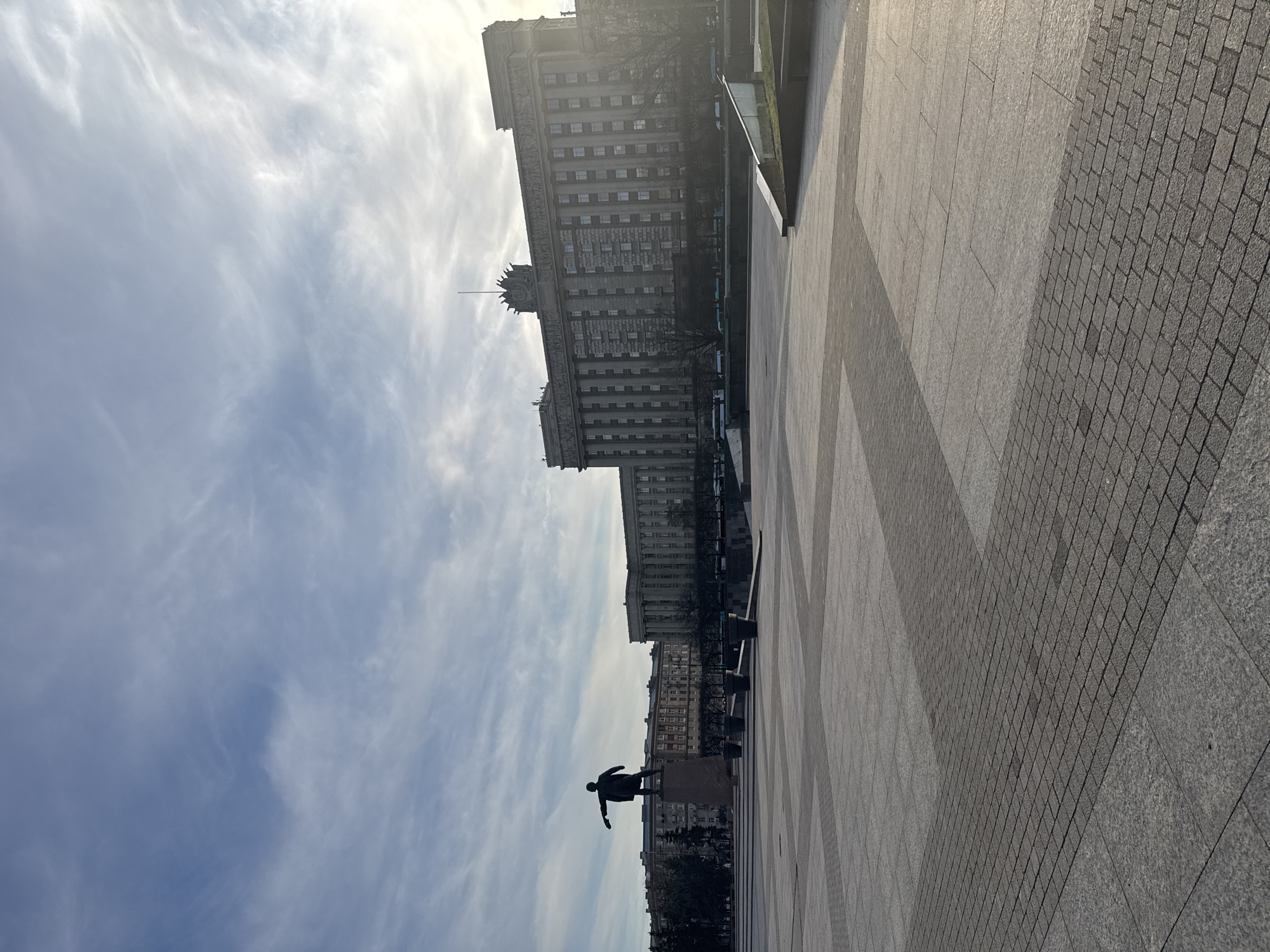 The image shows a wide-angle, long shot of a statue and a building in Saint Petersburg, Russia. The statue, likely a monument of Vladimir Lenin, stands on a tall pedestal. The building is a large, multi-story structure with a neoclassical architectural style and many windows. The foreground features a paved area with a combination of large stone slabs and smaller cobblestone sections. The sky is visible, with wispy clouds spread across a light blue background. The image has a slight upward angle, emphasizing the height of the building and statue.