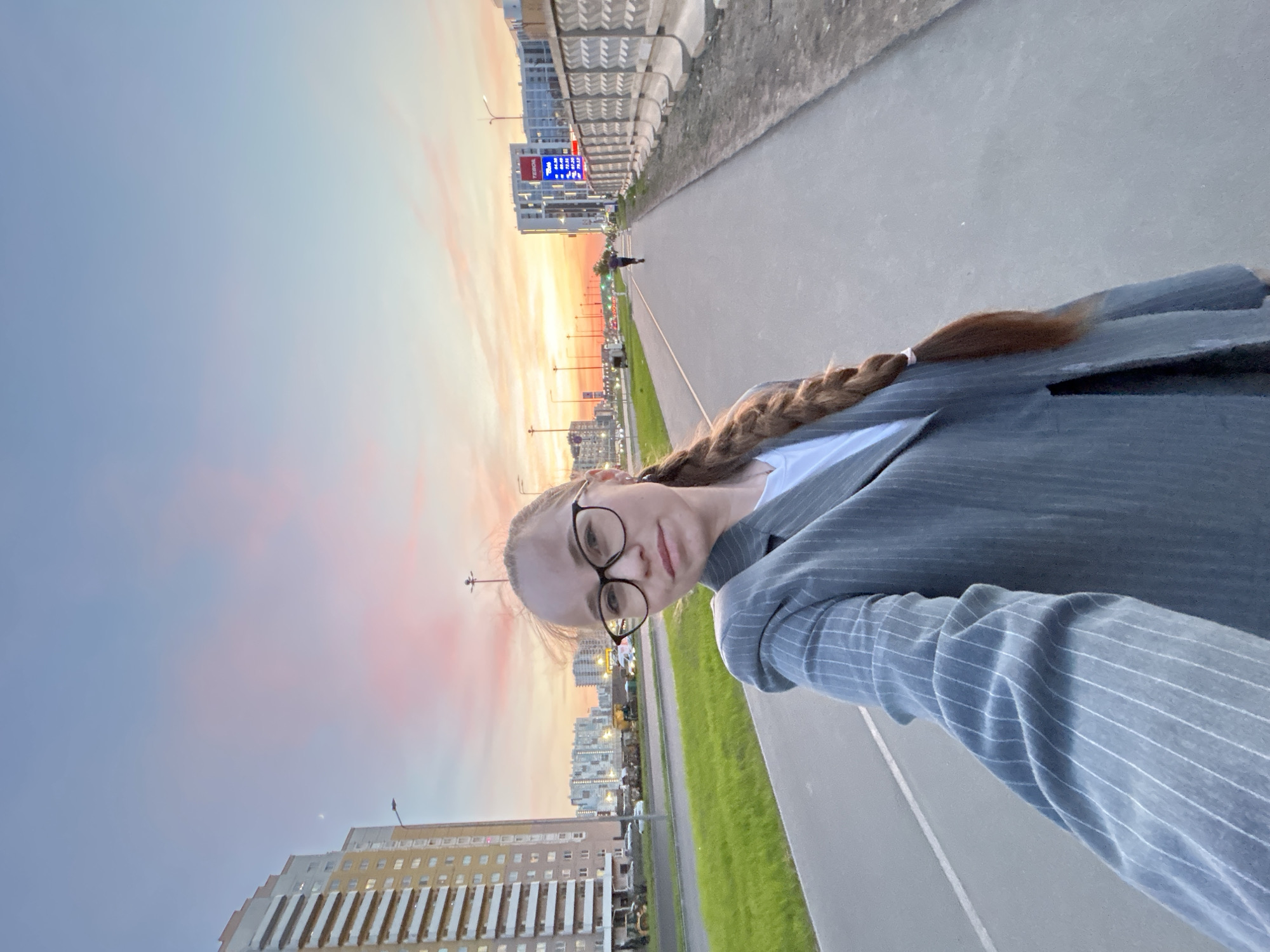 A selfie of a young woman with a braid, wearing glasses and a gray pinstripe blazer, taken at sunset in Saint Petersburg, Russia. The location is likely near the Lakhta Center, as the image includes modern high-rise buildings with distinctive architectural elements and the colors of a petrol station can be seen in the distance. A walkway or sidewalk is visible, with green grass on one side. A person is visible in the distance walking on the walkway. The sky is filled with colors of orange, pink and purple from the setting sun. The photo is taken at an angle.