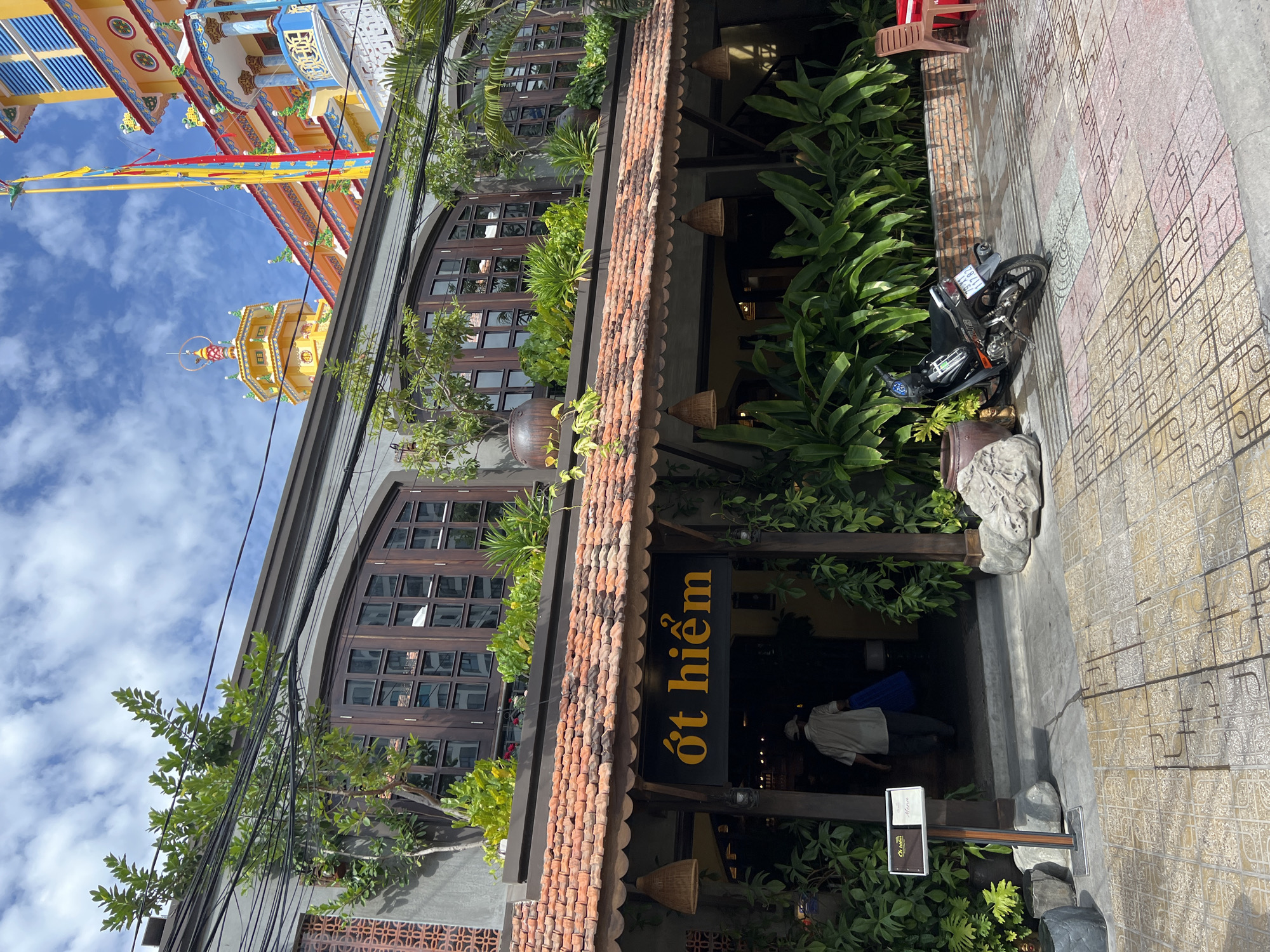 In Nha Trang, Vietnam, a street view captures a building entrance with a tiled roof and prominent signage reading "ớt hiểm" in yellow letters. Green foliage surrounds the entrance, with a person in a light-colored shirt and hat entering the establishment carrying a blue bag. A black motorcycle is parked on the patterned sidewalk to the right. In the background, a vibrant yellow and colorful structure, possibly a temple, rises against a cloudy blue sky, with various cables and wires crisscrossing overhead.