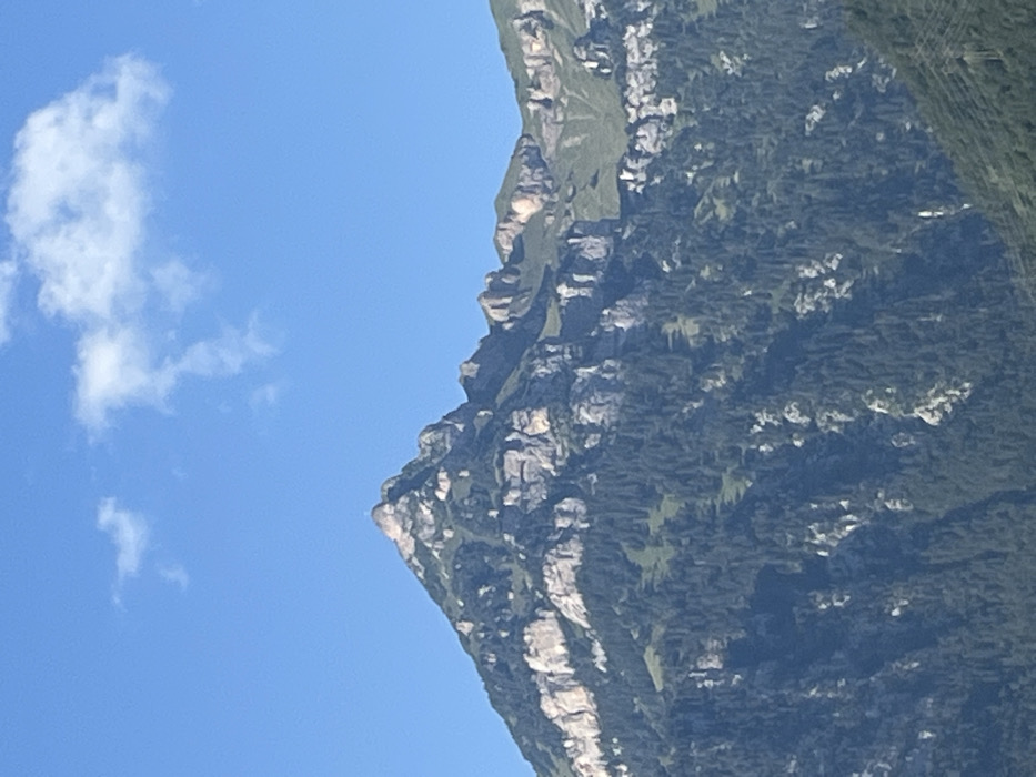 The image displays a prominent view of the Gross Windgällen mountain, observed from Seedorf (UR), Switzerland. The mountain's summit, situated centrally in the upper frame, is marked by a small, distinct cross. The upper and mid-slopes are characterized by a rugged terrain featuring large expanses of light-colored rock cliffs and outcrops, interspersed with areas of green alpine vegetation and dense, dark green coniferous forests that extend down the lower visible portions of the mountain. The sky above is a clear, bright blue with scattered, wispy white clouds. In the lower right corner, dark, angular lines consistent with a power line tower structure are partially visible. No human figures or activities are present.