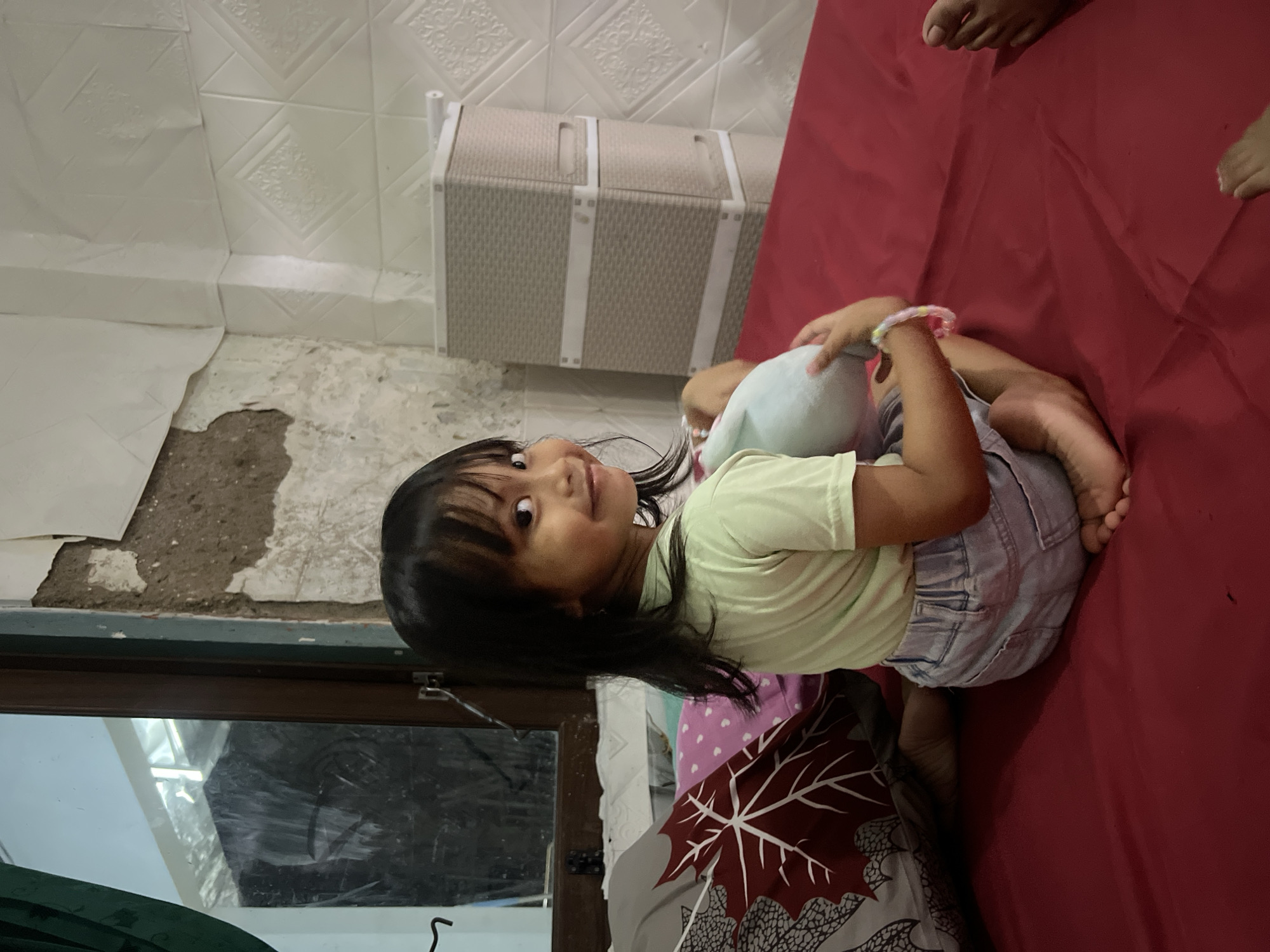 A young girl with dark hair is sitting on a red surface, likely a bed, in what appears to be a room in Kecamatan Pati, Indonesia. She is wearing a light green t-shirt and denim shorts, and is holding a light blue stuffed animal. Her gaze is directed towards the viewer with a slight smile. Behind her, a wall displays a rough, unfinished texture with peeling plaster, contrasting with a section covered in white, textured tiles. To the left, a window with a dark frame shows a reflection of what might be an outdoor scene or another room, partially obscured by a dark green curtain. Further to the right, stacked storage units with a woven texture are visible against the tiled wall. The overall impression is of an indoor domestic setting, potentially undergoing renovation or in a state of disrepair.