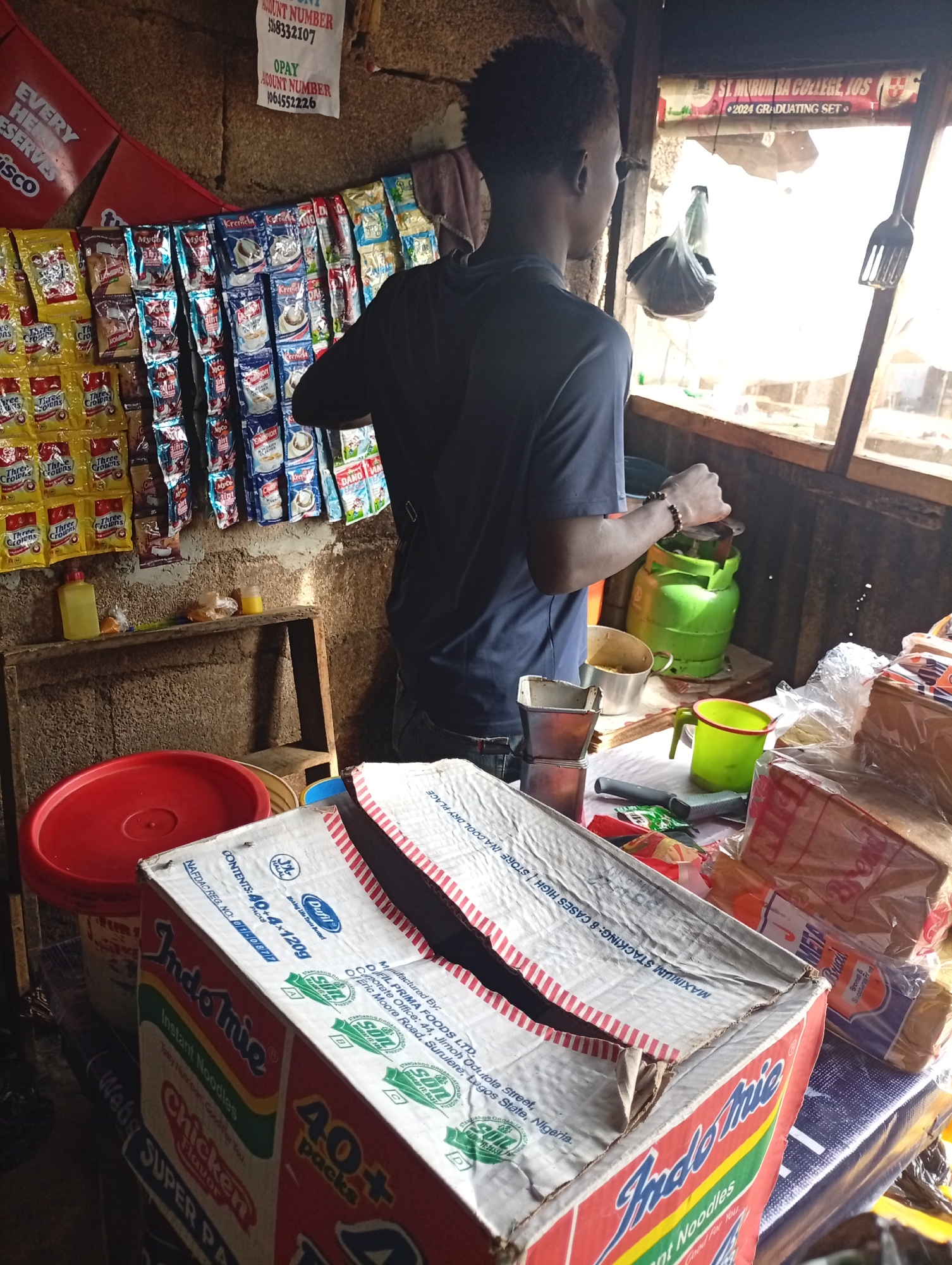 The image shows a young man standing in a small shop, possibly a convenience store or kiosk, in an urban setting. He is wearing a dark blue t-shirt and jeans. In front of him, there are shelves stocked with various food items, including packaged snacks and drinks. A green gas cylinder is visible on a counter. To the right, there is a window with a view of the outside, and a sign that reads "ST. MURUMBA COLLEGE, IOS 2024 GRADUATING SET". In the foreground, there are boxes of Indomie instant noodles and other packaged goods. The environment appears rustic and somewhat cluttered, suggesting a local, everyday business.