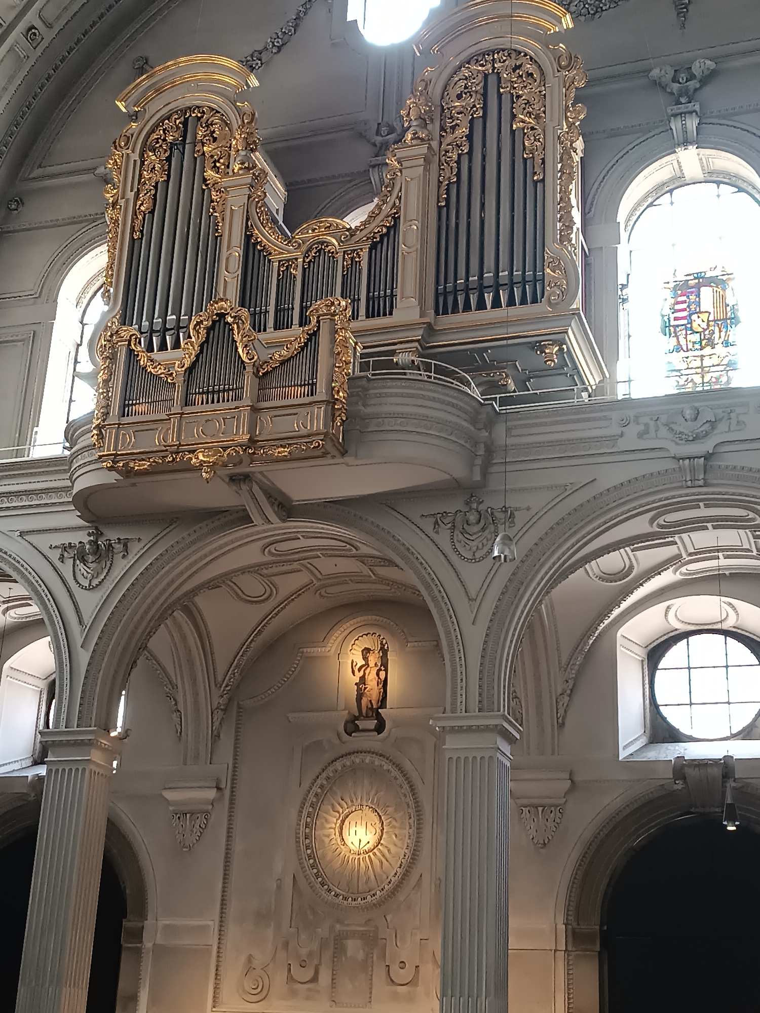 The image depicts the interior of a grand church in Munich, Germany, specifically the St. Michael's Church. The dominant feature is a large, ornate pipe organ situated high on the wall, adorned with intricate gold detailing. Below the organ, the church's architecture is visible, characterized by arched doorways, decorative moldings, and large windows, including a circular window to the right. A stained-glass window with a coat of arms is partially visible through one of the arched windows on the right side of the image. In the center of the lower section, there is a niche containing a statue of a religious figure, and below that, a circular decorative element with the letters "IHS" inscribed within radiating lines. The lighting suggests natural light entering from the windows, creating a bright, airy atmosphere within the church. The scene is static and devoid of any people.