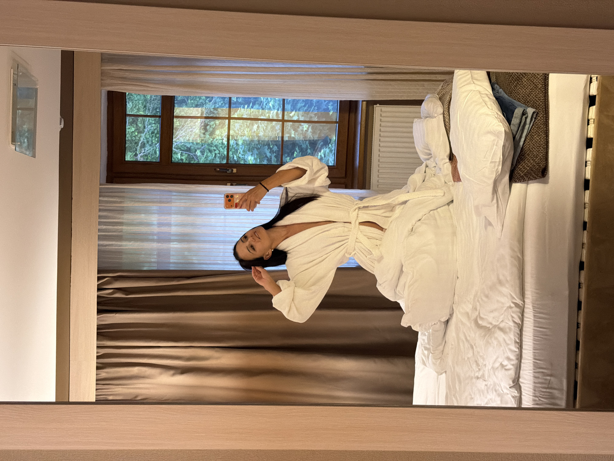 A full-length mirror selfie depicts a woman sitting on a bed in a room in Svetlogorsk, Russia. The woman, with long dark hair, wears an open white terrycloth bathrobe and holds an orange-cased smartphone in her right hand, angled towards the mirror. Her left hand is raised towards her head. She sits on a bed with white bedding, featuring a rumpled duvet and pillows. Folded clothing, including blue jeans and a brown patterned item, rests on a pillow on the bed's right side. Behind the woman, a multi-pane wooden-framed window reveals dense green foliage outside. Light brown opaque curtains cover the left side of the window, while sheer white curtains are on the right. A white radiator is situated below the window. The room's ceiling is white, with a reflective rectangular fixture in the upper right, and a dark brown trim runs across the upper wall, beneath which a lighter wooden beam is visible. The mirror is framed by light-colored wood on both sides.