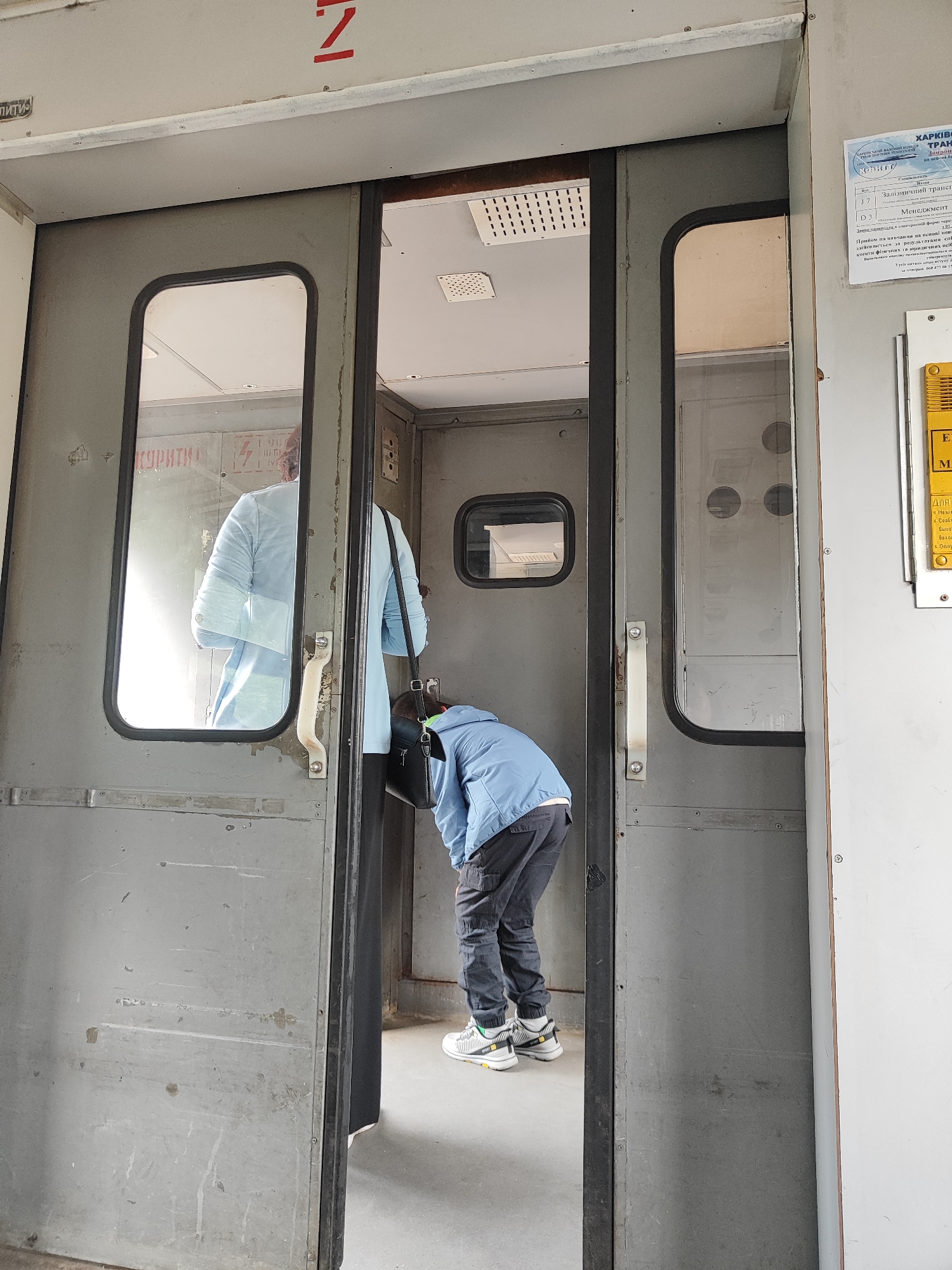 The image shows an interior view of a train car or similar transportation vehicle, with a double door in the foreground. On the left, a person in a light blue jacket is reflected in a window and appears to be standing. On the right, a person wearing a blue jacket and dark pants is bent over, possibly looking for something on the floor. They are wearing athletic shoes. The environment appears utilitarian, with metal doors and walls. There are signs on the wall to the right, one of which has Ukrainian text. The overall impression is one of a functional, possibly older, public transportation setting.