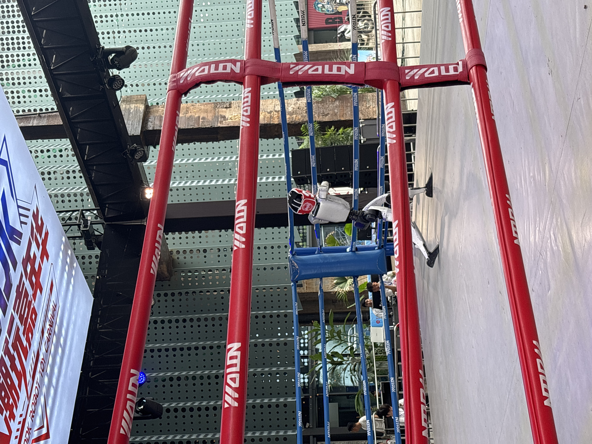 The image shows a robot in a boxing ring, potentially at an event called "Dook Robot Toy Carnival" in Dadukou, China. The robot, which is white and red with a helmet, is in a boxing stance. The boxing ring has red ropes and blue padding. The background shows a building with a perforated facade and a banner overhead with Chinese characters and the text "Dook Robot Toy Carnival." Several people are visible in the background, some spectating and others near the ring. The environment appears to be outdoors or in a large open space, with some greenery visible.
