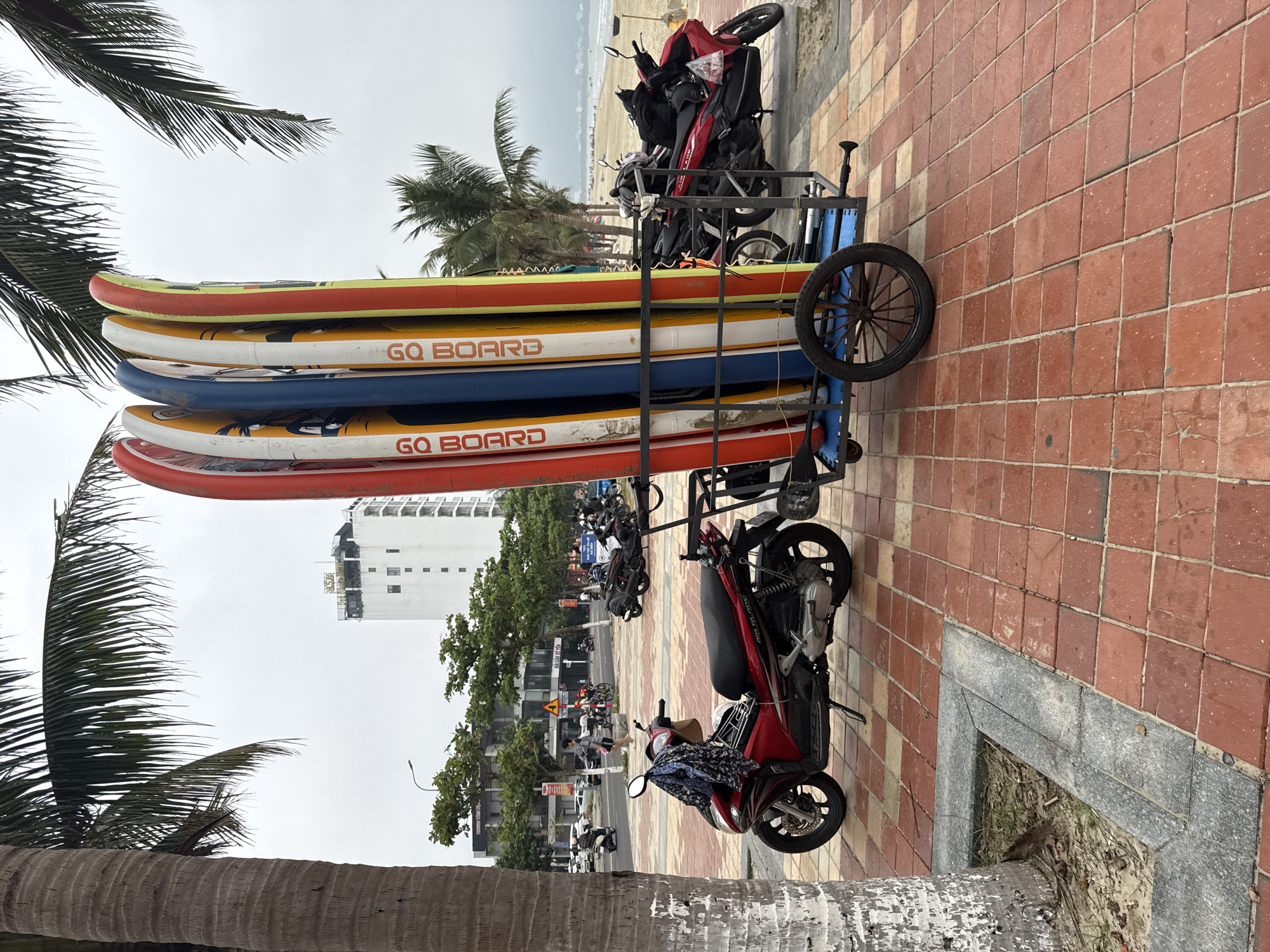The image captures a coastal street scene in Đà Nẵng, Vietnam, specifically along a boulevard adjacent to a beach, likely My Khe Beach. In the foreground, a reddish-brown tiled sidewalk features the base of a large palm tree on the left. A red and black Honda Air Blade scooter is parked on its kickstand, with a patterned cloth and a plastic bag draped over its handlebars. To the right, a black metal utility cart with a blue base and two large wooden-spoked wheels holds seven distinctively colored stand-up paddleboards or surfboards vertically. These boards are predominantly red, white, yellow, and blue, each prominently marked "GO BOARD".
In the mid-ground, a street runs parallel to the sidewalk, lined with various green trees and numerous parked scooters. A single individual is discernible walking on the sidewalk, wearing a light-colored shirt and shorts. Other vehicles and distant figures are visible further down the street. The background features a tall, white multi-story building, identifiable as "HOÀNG SA" by a sign on its rooftop. To the right, a sandy beach extends towards the ocean under an overcast sky, with faint outlines of additional structures or people along the distant shoreline.