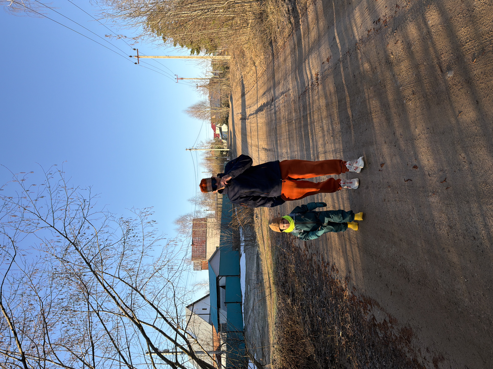 The image shows two people standing on a dirt road in Chaikovsky, Russia. The road is lined with trees and power lines. A building is visible in the background. The first person is wearing an orange hat, a dark jacket, and orange pants, and white sneakers. The second person is wearing a green jacket, light brown pants, and yellow boots. The sky is blue and the weather appears clear.