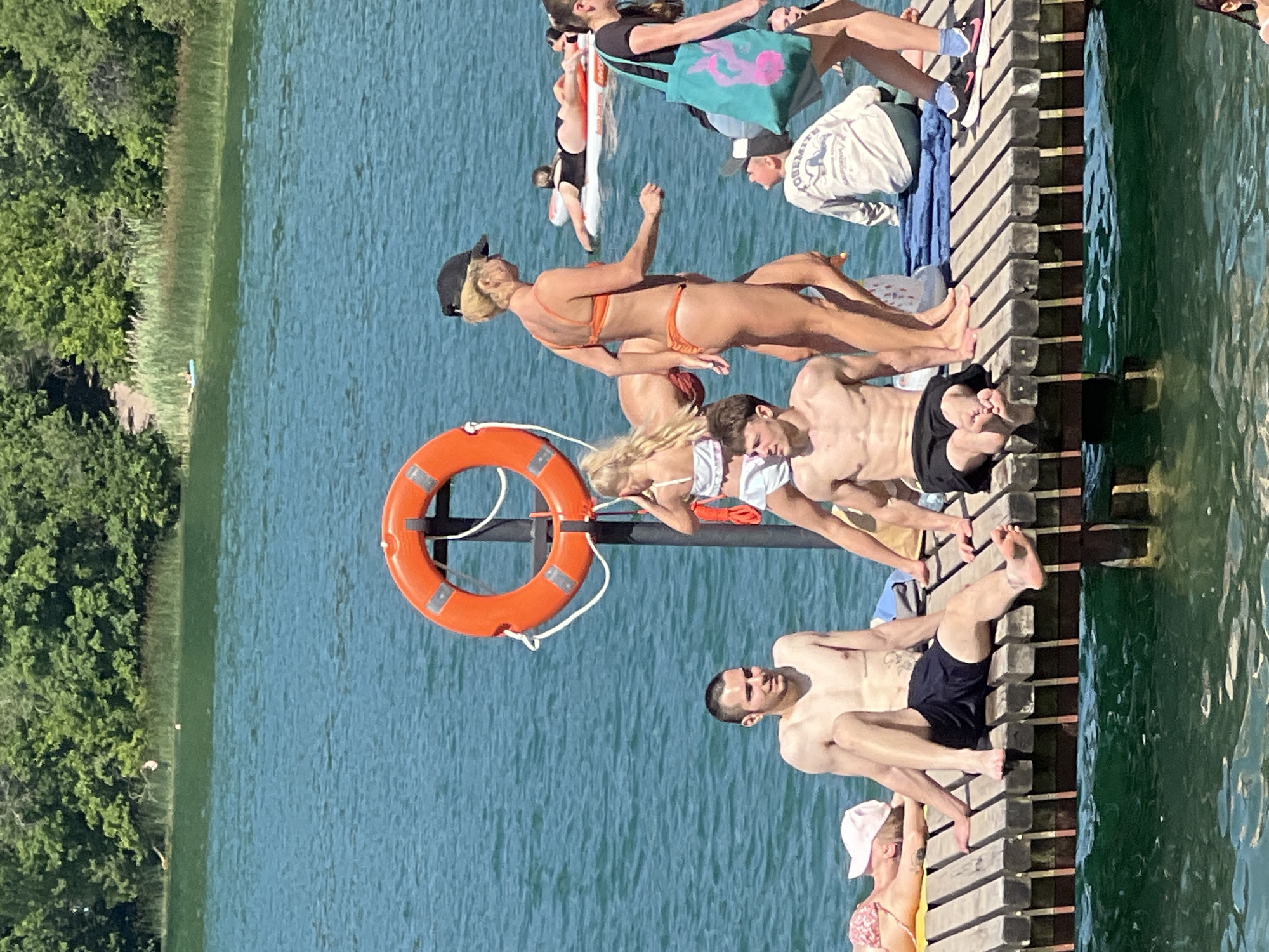 The image depicts a group of people enjoying a sunny day at a lake in Vilnius, Lithuania. They are gathered on a wooden dock, with the clear blue water of the lake providing a refreshing backdrop. The scene is bustling with activity as individuals are sunbathing, lounging, and interacting with each other.
The subjects are diverse, including men, women, and possibly teenagers. They are dressed in swimwear, casual summer attire, and some are wearing hats. One prominent object is an orange life preserver attached to the dock, suggesting it is a recreational area. The people are positioned in various poses: some are sitting or lying down, others are standing or leaning.
The atmosphere appears relaxed and joyful, typical of a summer outing. The bright sunlight indicates it is daytime, likely midday or early afternoon, and the weather seems warm and pleasant. The lush green trees on the shoreline provide a natural setting for the lake.
One person on the dock is wearing a white long-sleeved shirt with visible text that reads "YOSEMITE CALIFORNIA". Another person is holding a turquoise bag with a pink octopus-like graphic. The overall impression is one of leisure and fun, with people taking advantage of the beautiful weather and water to relax and socialize.