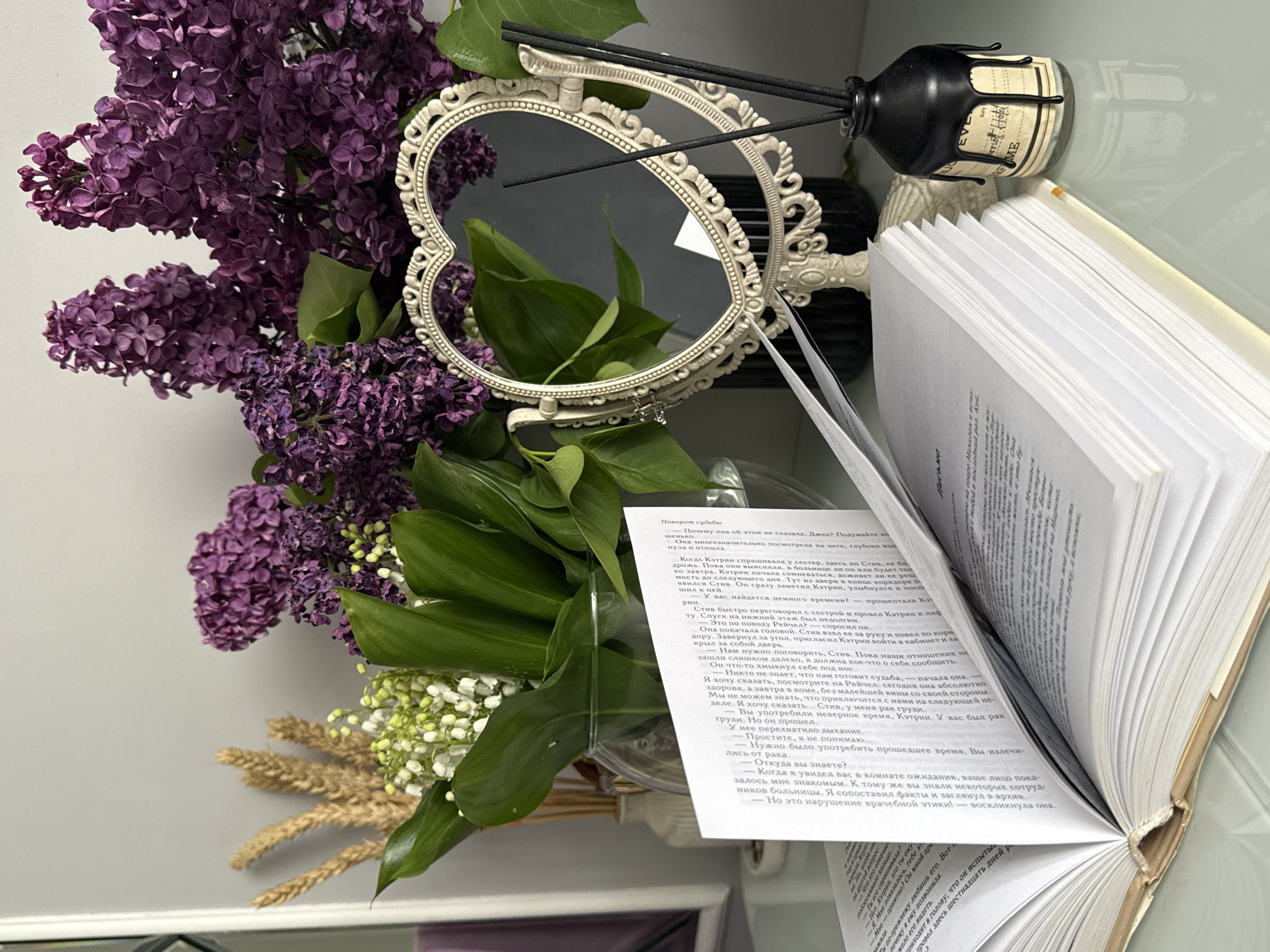 The image captures an indoor still life arrangement featuring a bouquet of flowers, an ornate mirror, an open book, and a diffuser bottle, set on a light-colored surface against a plain background.
The bouquet is a central element, comprising vibrant clusters of deep purple lilac blossoms, delicate white lily of the valley, and several stalks of dried golden wheat, lending a rustic yet elegant feel to the display. Positioned behind the bouquet is a vintage-style, heart-shaped mirror with an intricate, light-colored frame, reflecting parts of the room and some green foliage.
To the right of the mirror, an open book with numerous pages is prominently displayed, revealing text written in Russian. Adjacent to the book and behind the mirror is a distinctive black diffuser bottle, designed with a "dripping wax" effect around its neck. Several black reeds are inserted into the bottle.
No people are visible in the scene. The lighting appears soft and ambient, creating a serene and inviting atmosphere.
Visible text on the book's pages includes the title "Поворот судьбы" (Turn of fate) and dialogue between characters named Jack, Catherine, Steve, and Rachel. The conversation delves into serious topics such as a character having "рак груди" (breast cancer) and a "нарушение врачебной этики!" (violation of medical ethics!). The label on the diffuser bottle clearly displays the words "EVE" and "Key", with other fragmented text like "ME".
