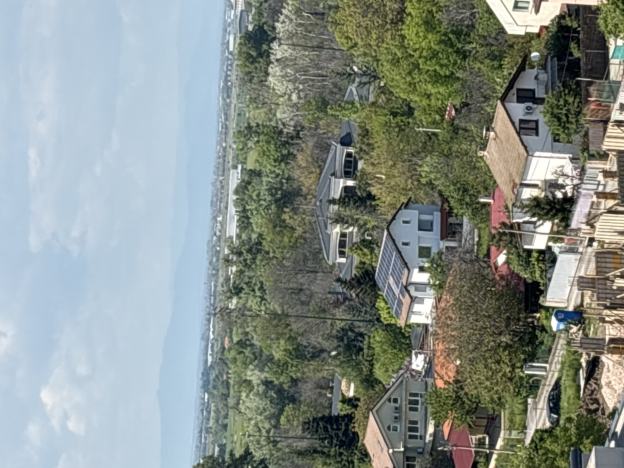 This image captures a residential area in Plovdiv, Bulgaria, seen from an elevated vantage point. The scene is dominated by closely packed houses with varying roof colors (red, brown, and white) surrounded by lush green trees. Some houses have visible solar panels on their roofs. In the distance, a hazy landscape of plains or fields meets the sky. A road or highway can be seen running through the lower portion of the image.