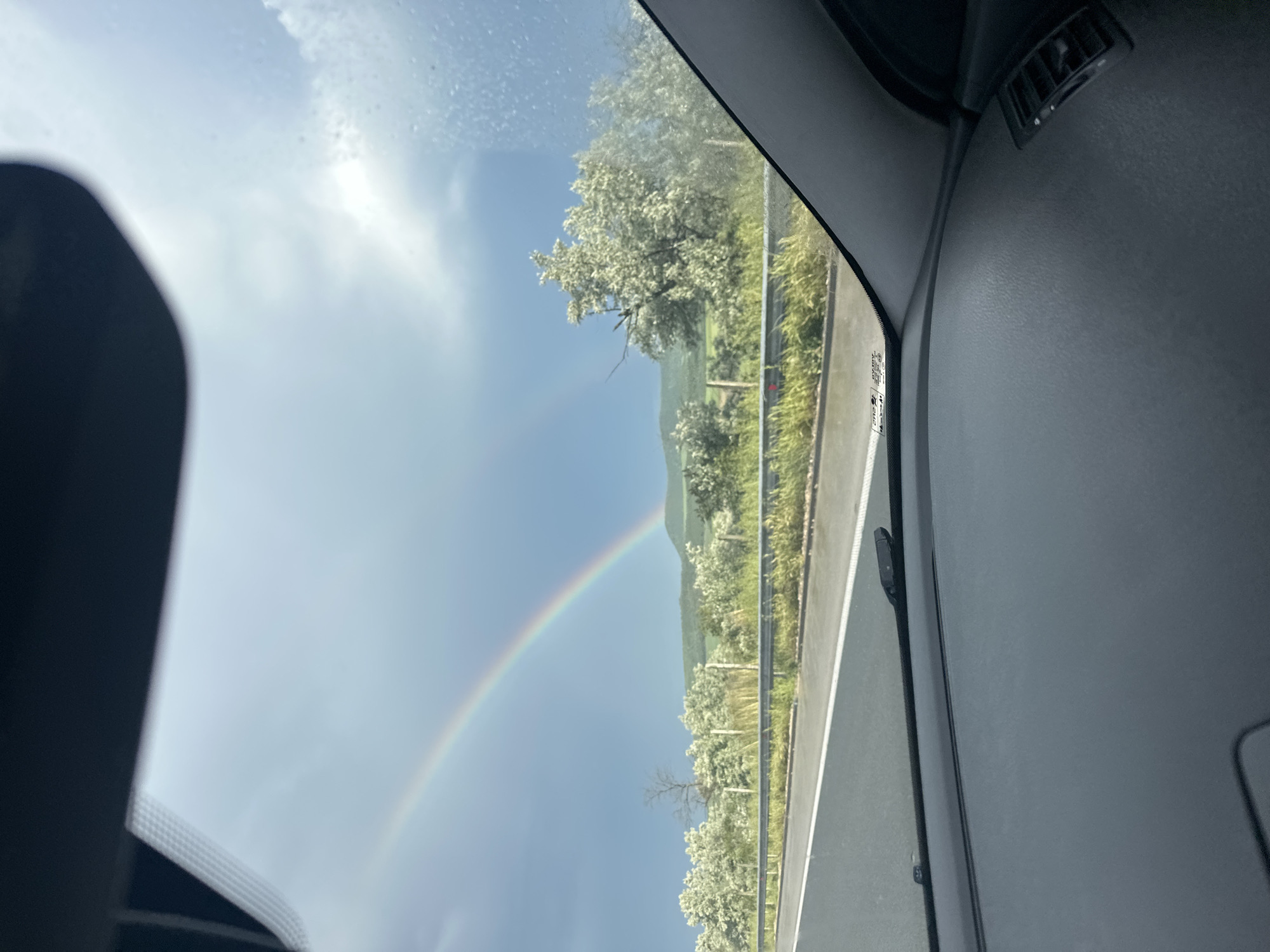 The image, taken from inside a vehicle, shows a partial rainbow in the sky above a highway and a wooded hillside. The car's interior, including the dashboard and a side window frame, are visible. The photograph appears to be taken while moving on the highway. The surrounding environment is green, consisting of trees and vegetation along the highway's edge. The location cannot be determined from the image alone.