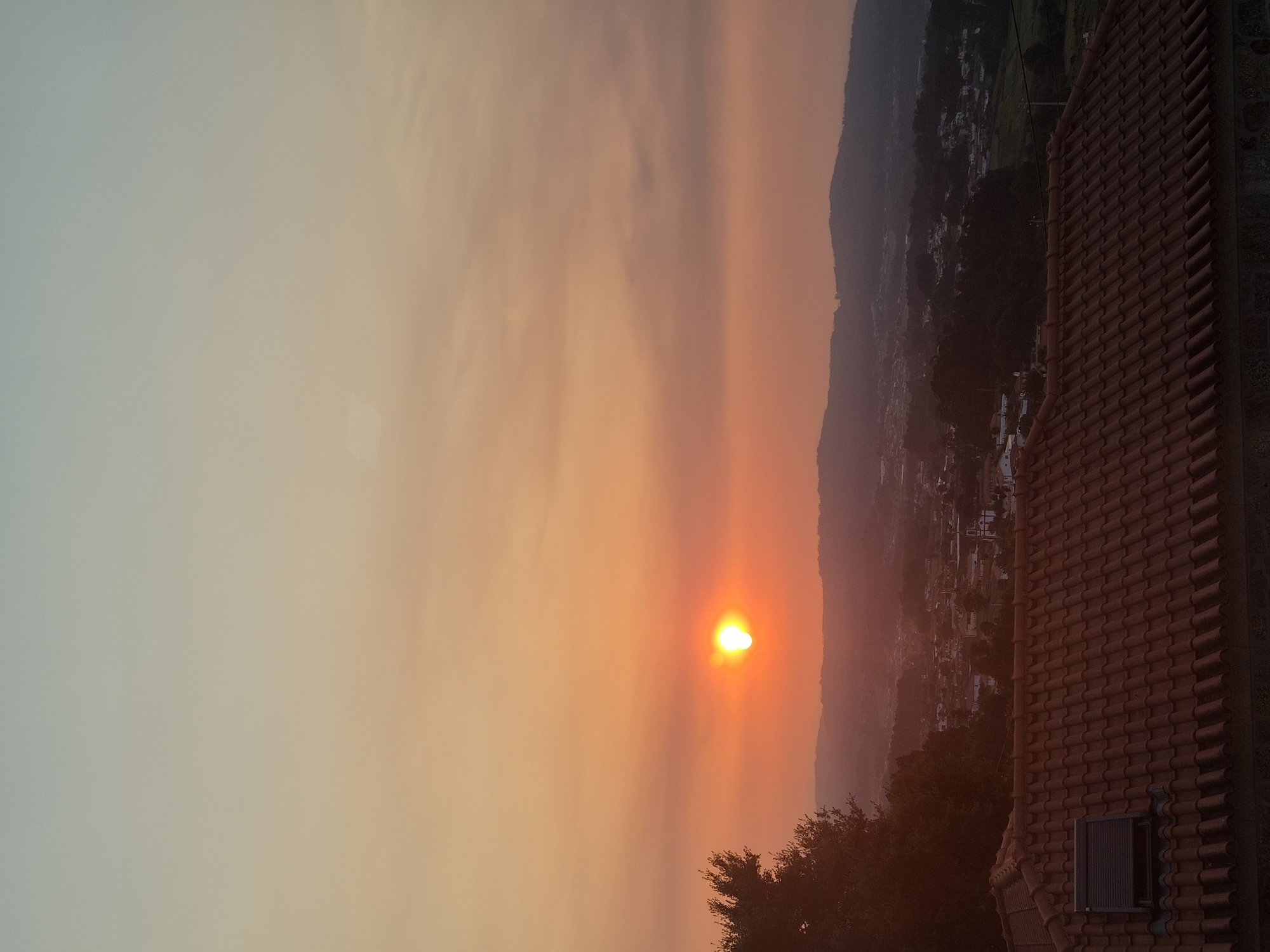 The image depicts a sunset over the city of Barcelos, Portugal. The sun, a bright orb with a halo, is positioned in the center-left of the frame, casting a warm, orange glow across the sky and the landscape below. The sky transitions from a pale blue at the top to a rich orange near the horizon, with wispy clouds visible.
In the foreground, the reddish-brown tiled roof of a building dominates the lower portion of the image. The roof has a weathered appearance and features a dark, rectangular skylight with a rolled-down shutter. To the left of the roof, a dense, dark green tree with textured foliage extends into the frame from the edge.
Beyond the immediate foreground, a valley unfolds, revealing the cityscape of Barcelos. Houses with white and terracotta roofs are nestled among rolling hills and dense vegetation. Distant, hazy mountains form the backdrop, their silhouettes softened by the sunset's light. The overall scene conveys a sense of tranquility and the beauty of a rural European sunset.