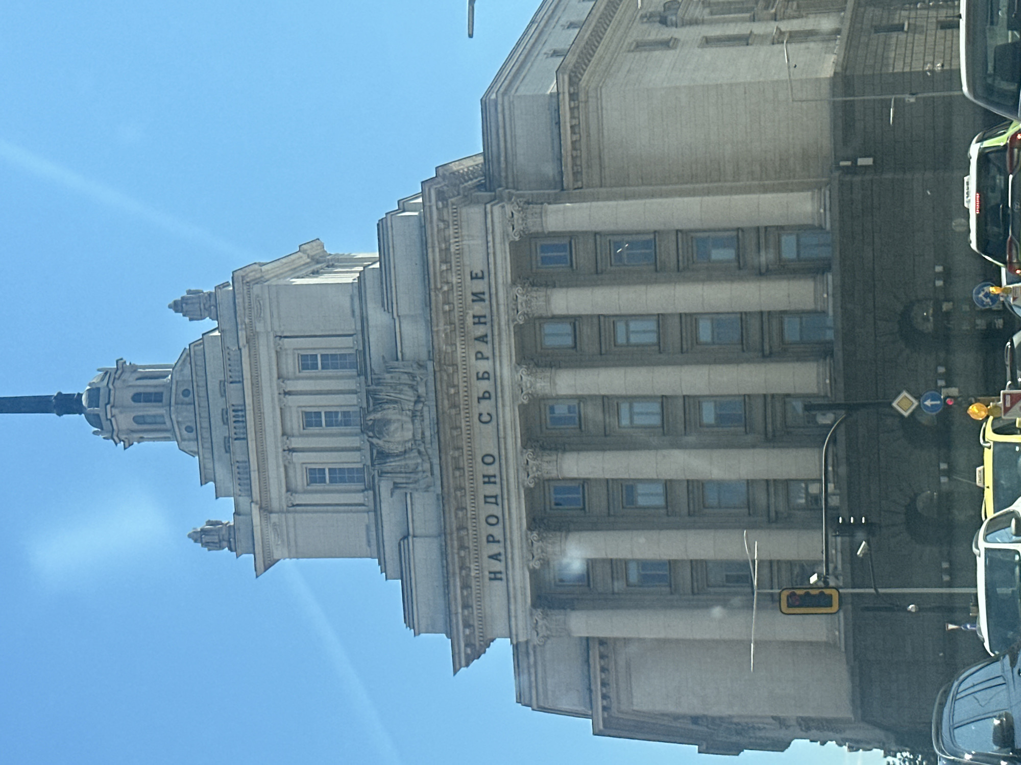 The image shows the National Assembly of Bulgaria building in Sofia, Bulgaria. It is a large, multi-story building with a classical architectural style, featuring columns, windows, and decorative elements. The facade has "НАРОДНО СЪБРАНИЕ" (National Assembly) inscribed vertically in Cyrillic letters. The building's tower is visible against a clear blue sky. Street lights and vehicular traffic are visible on the right, suggesting a city setting.