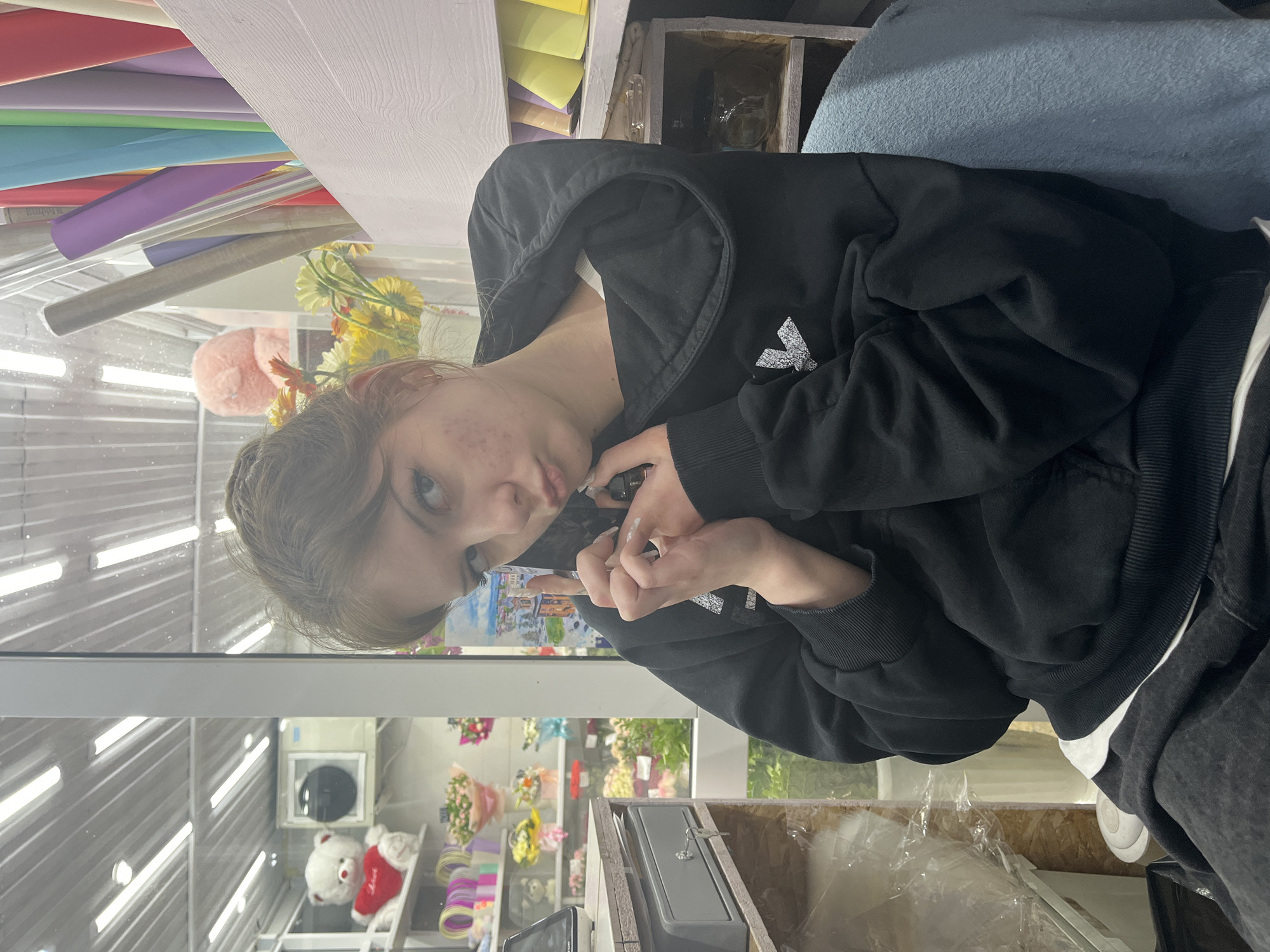 A person wearing a black hooded sweatshirt is holding a cell phone to their ear. They are standing in an indoor space with an overhead lighting structure and a display case behind them containing floral arrangements and a teddy bear. The background includes colorful rolls of paper and some shelves with additional items. The location is Unknown, Unknown.