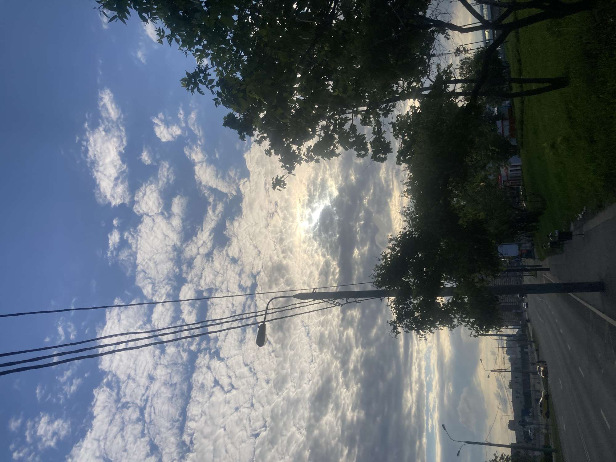 The image depicts a street scene in Moscow, Russia. The perspective is from ground level, looking upward at an angle. The sky is predominantly blue with scattered altocumulus clouds, and the sun is partially obscured by the clouds. Overhead power lines are visible, along with a street light. Trees line the street, their foliage partially obscuring the view. A road is visible with light traffic. Buildings are visible in the distance along the road. A grassy area is present to the right of the road. The photo appears to be taken in the late afternoon or early evening.