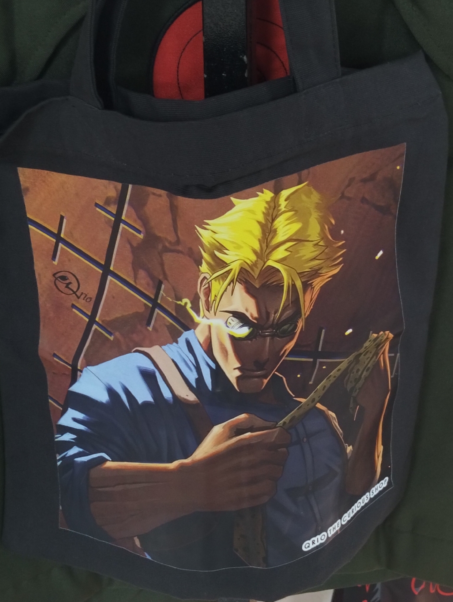 A black tote bag with a printed anime-style illustration of a male character with spiky blonde hair, wearing glasses and a blue shirt, is displayed. The character is holding a thin, segmented object, possibly a twig or a piece of wood. The background of the illustration features a brown, grid-like pattern with diagonal lines. A logo that appears to be "QriO" with an eye symbol is visible on the left side of the illustration, and text reading "QriO THE CURIOUS SHOP" is present on the bottom right. The tote bag is partially folded and placed against a backdrop of green fabric. The image appears to have been taken indoors, possibly in a shop or market in Dhaka, Bangladesh, as suggested by the context of a previous query.
